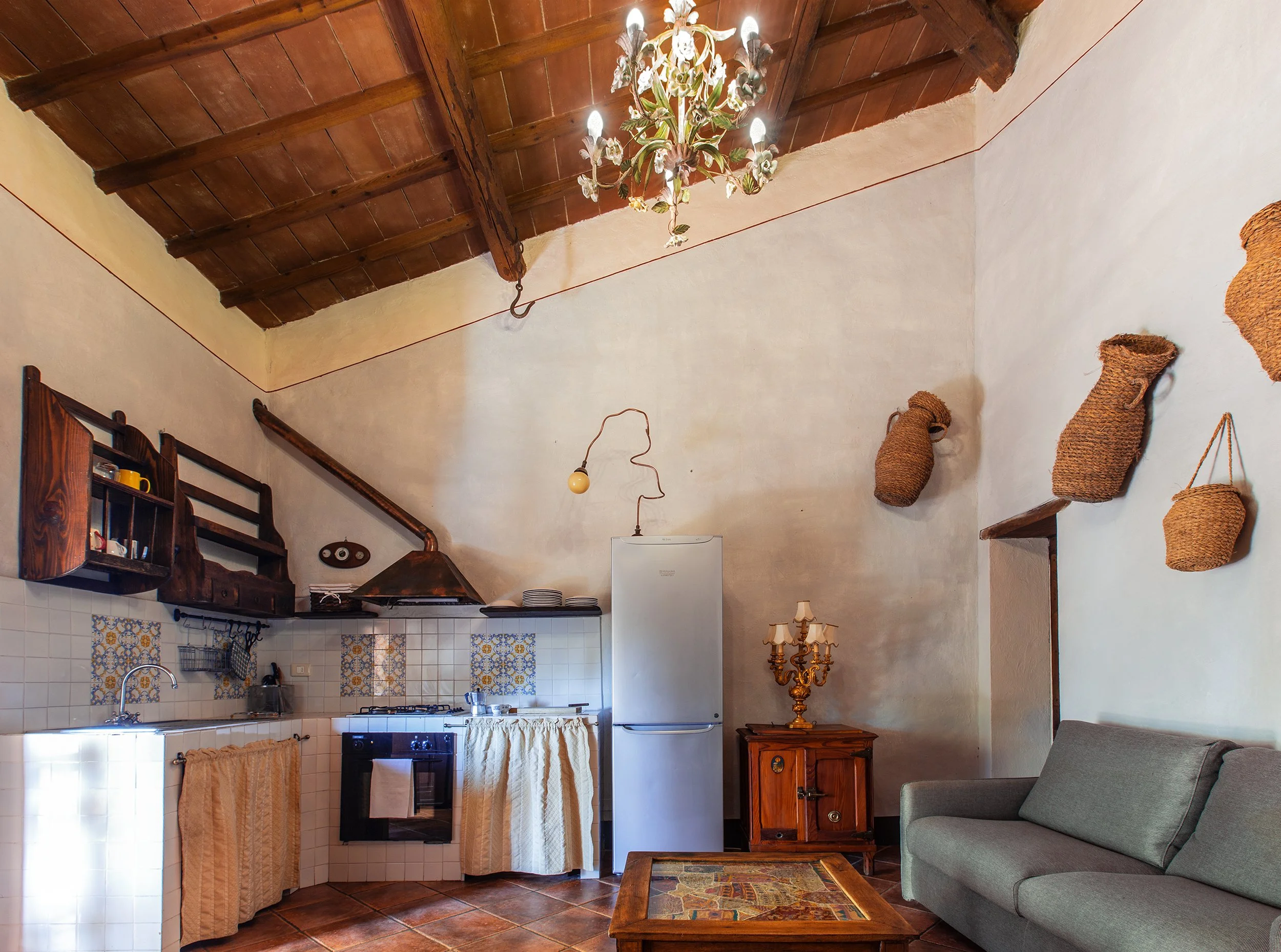 Interno di una cucina in stile rustico. Pareti bianche con decorazioni in ceramica e oggetti in vimini appesi alle pareti. Parete superiore con travi in legno a vista, soffitto in legno e lampadario decorativo. Cucina con forno a gas, frigorifero e l