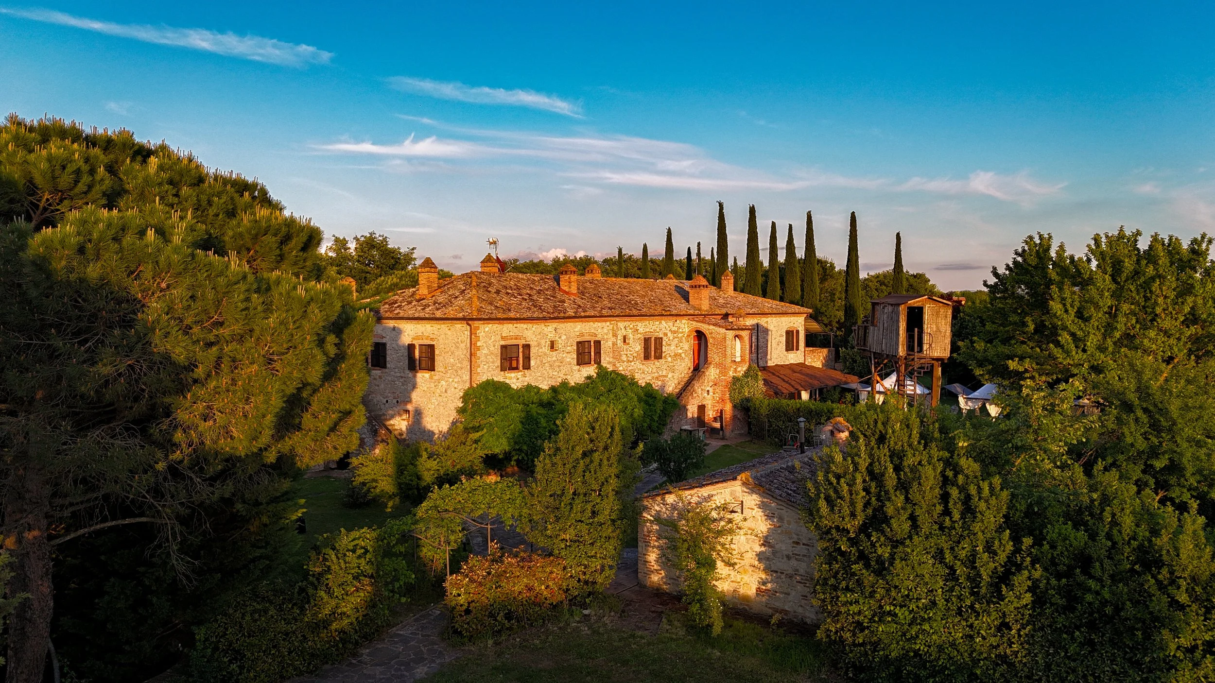 Un edificio in pietra circondato da alberi verdi, sotto un cielo blu con qualche nuvola, al tramonto.