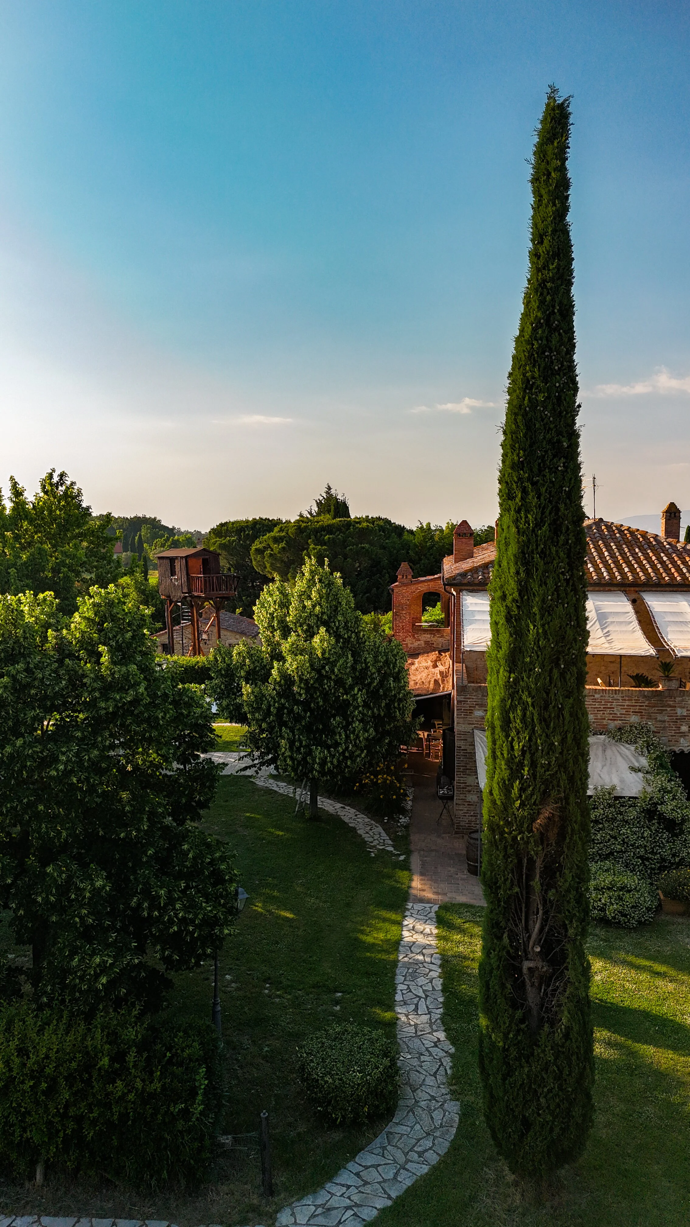 Un giardino con un sentiero di pietre che attraversa erba e alberi, con una casa in mattoni sullo sfondo e un cielo sereno sopra.