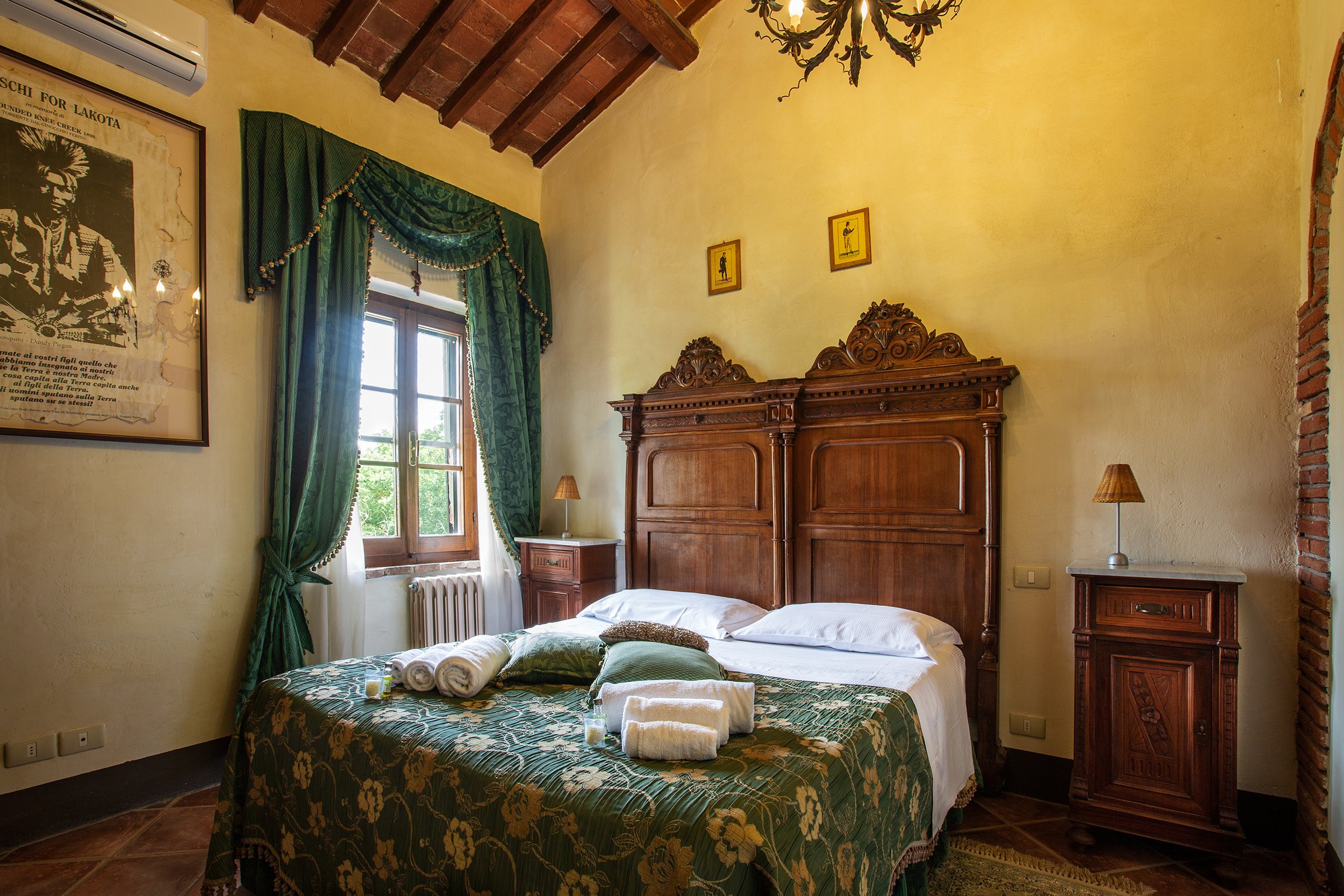 Camera da letto con pareti gialle, letto matrimoniale con copriletto verde con motivo floreale, letto con coperte piegate e asciugamani, armadio di legno intagliato, finestra con tende verde e stesse tonalità, lampade da comodino in tessuto, quadro con immagine di un personaggio e testo, parete in pietra a vista. Ceilings con travi di legno e lampadario decorativo.