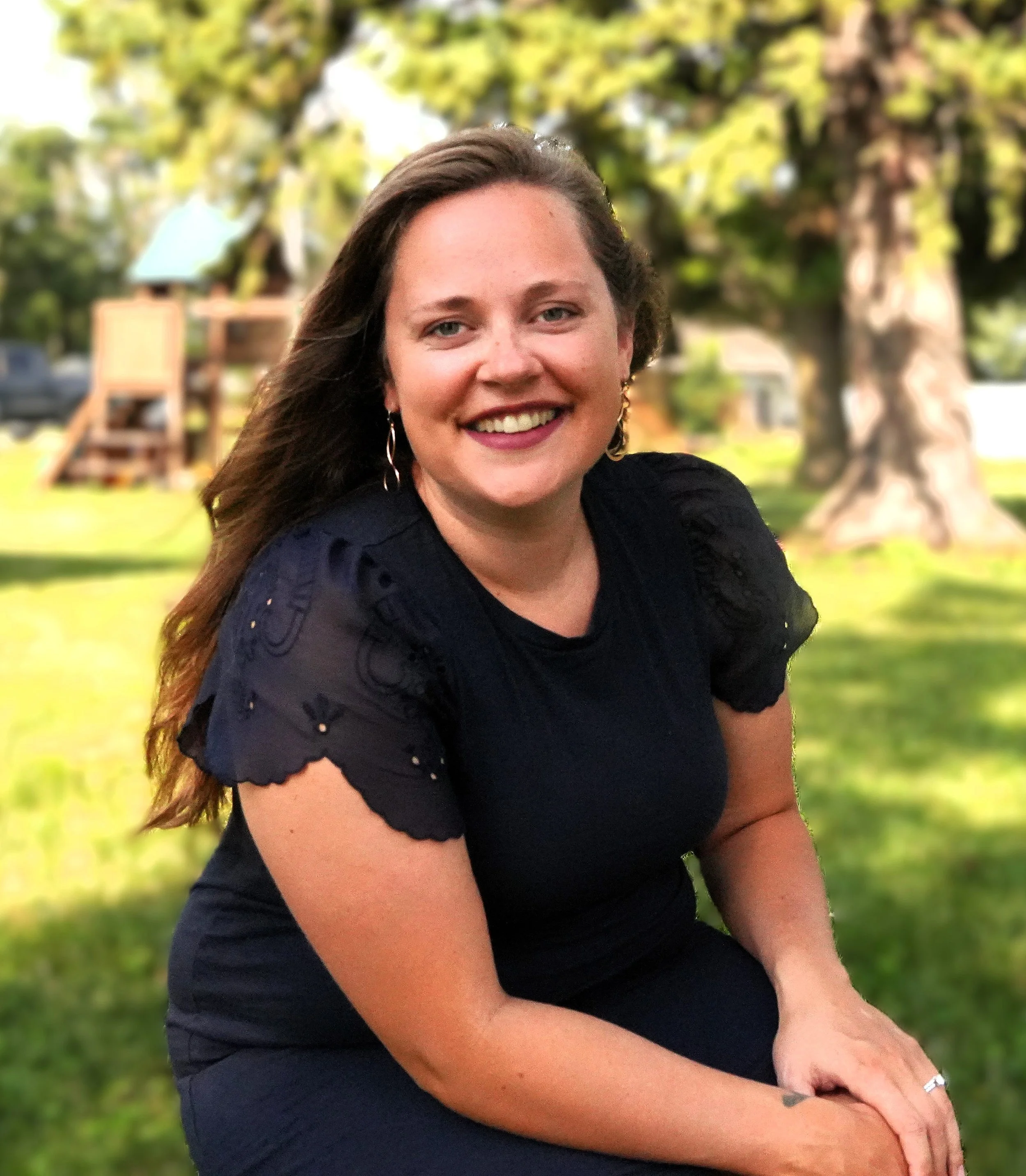 Smiling woman with long brown hair wearing a black top, sitting outdoors on grass with trees and a wooden play structure in the background.