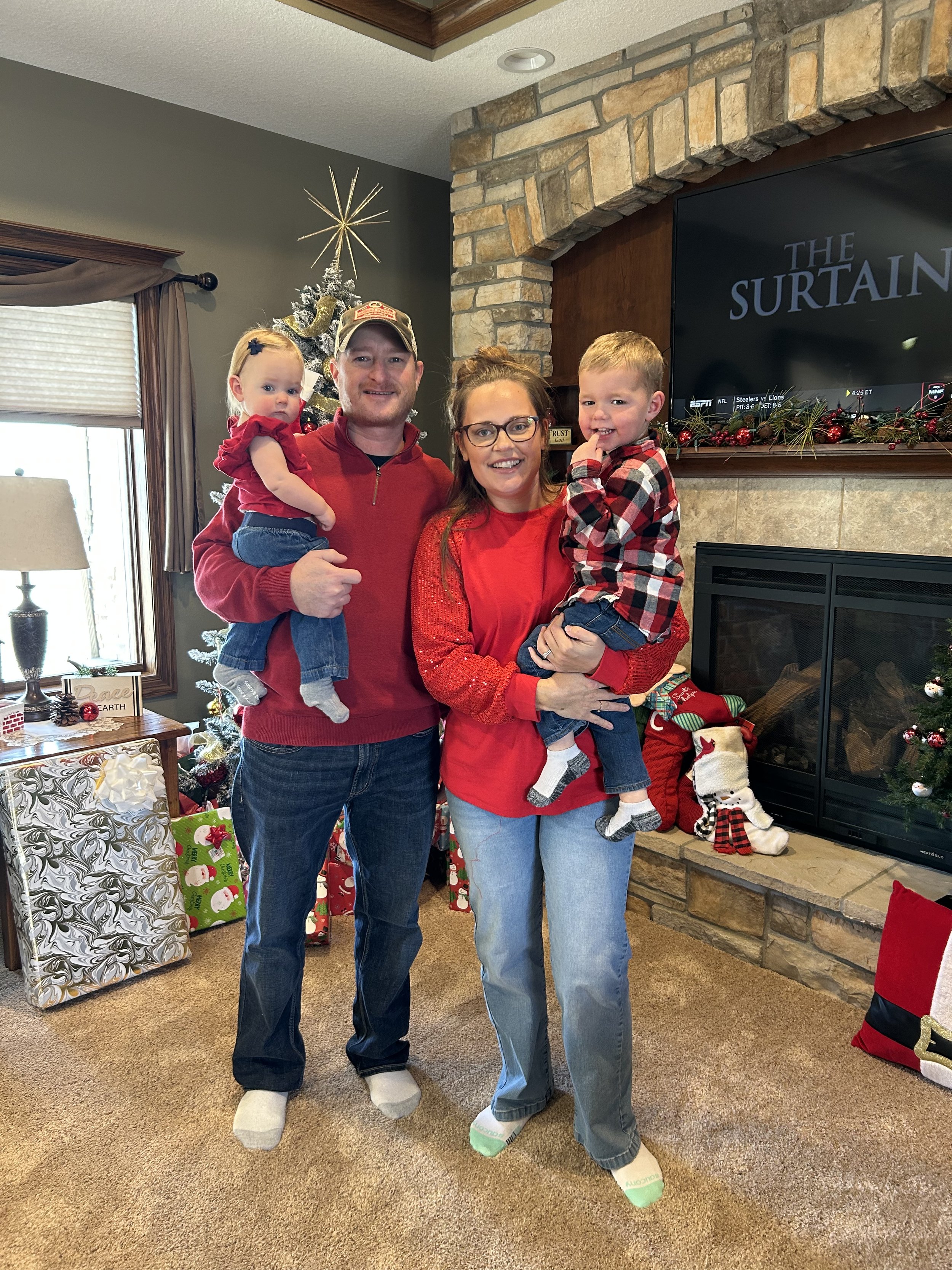 A family of four, wearing socks, standing in a living room decorated for Christmas, in front of a fireplace and Christmas stockings. They are smiling and posing for the camera.