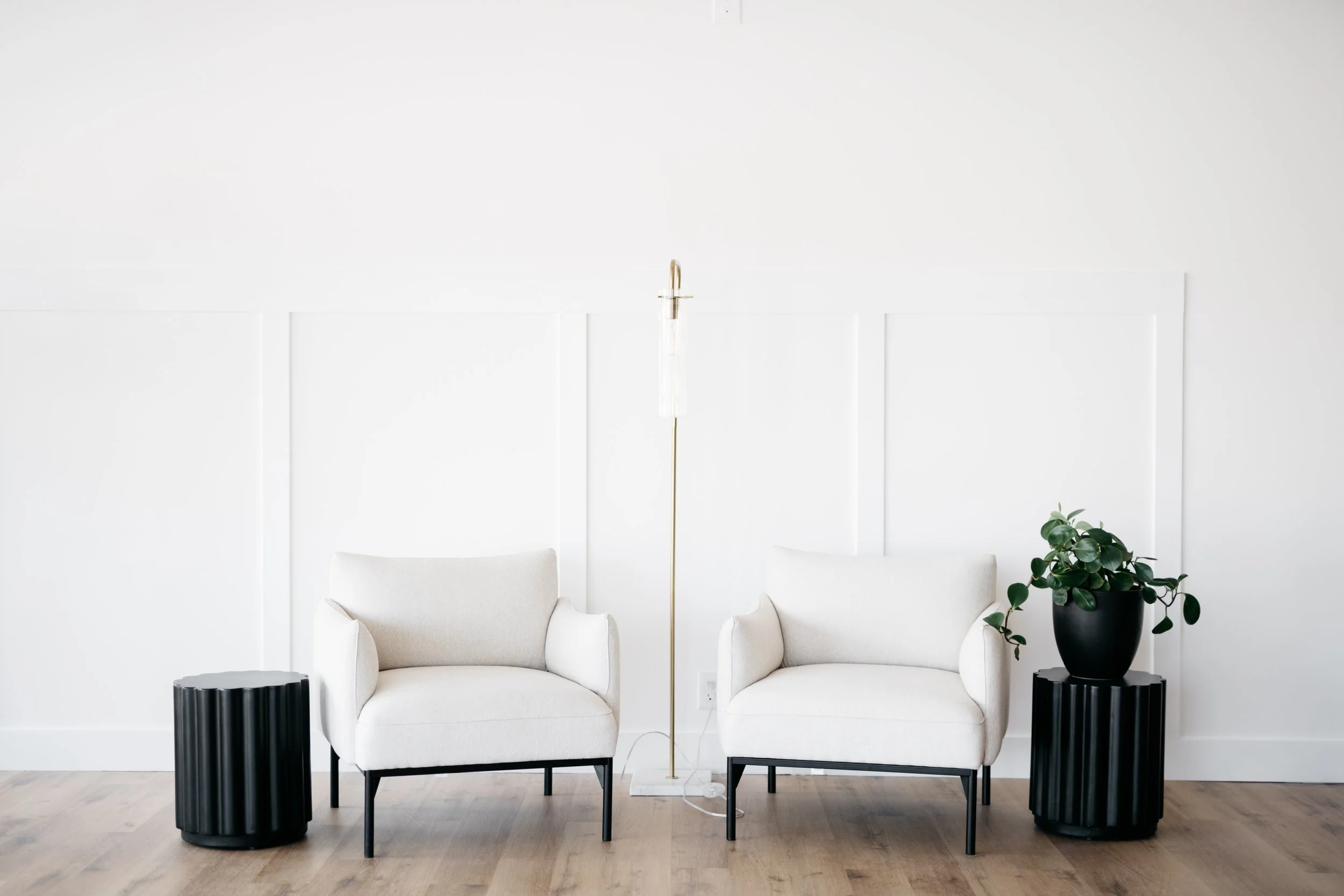 Minimalist white living room set with two white chairs, black side tables, a gold floor lamp, and live potted plants.