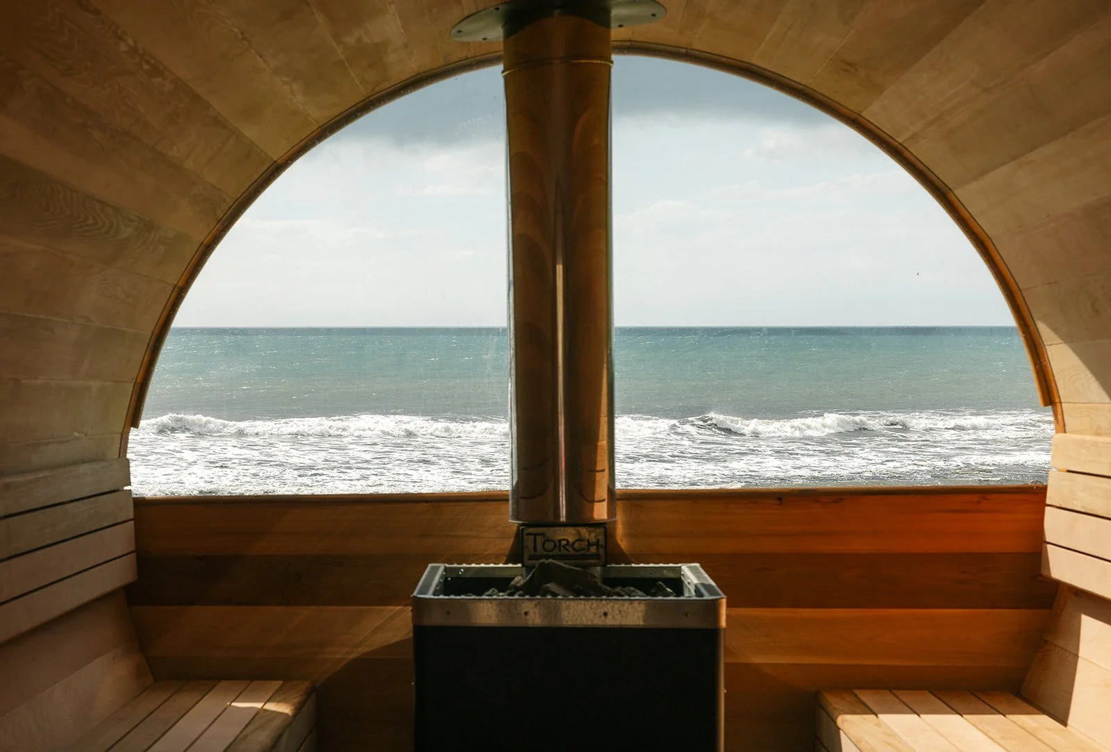 Inside a wooden sauna with a large arched window overlooking the ocean and waves.