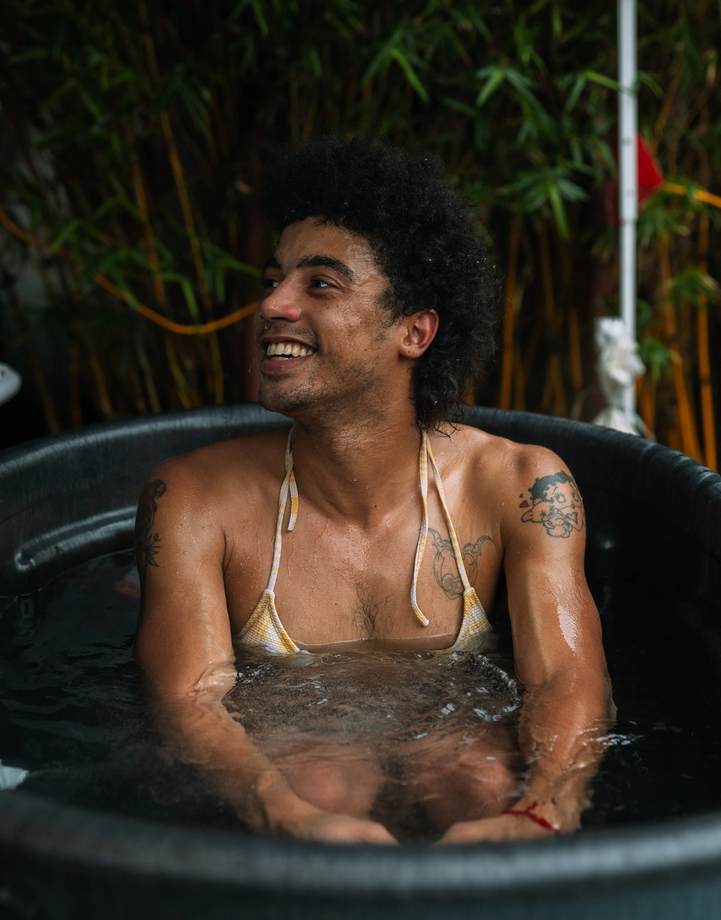 A woman with dark curly hair and tattoos smiling while sitting in a hot tub with water, with green foliage in the background.