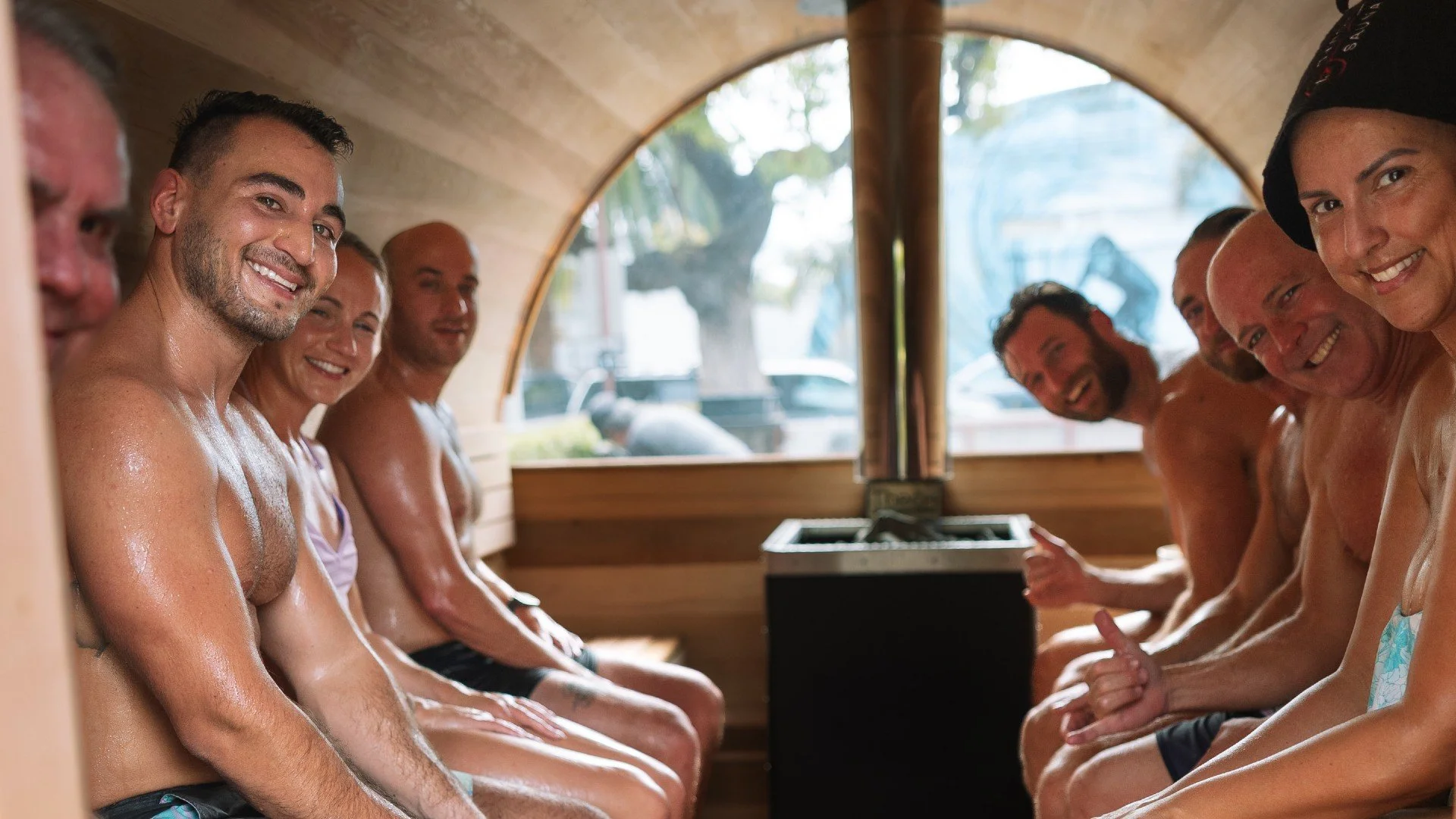 A group of smiling people, mostly men, sitting topless inside a sauna, with a wooden interior and a window showing an outdoor scene.