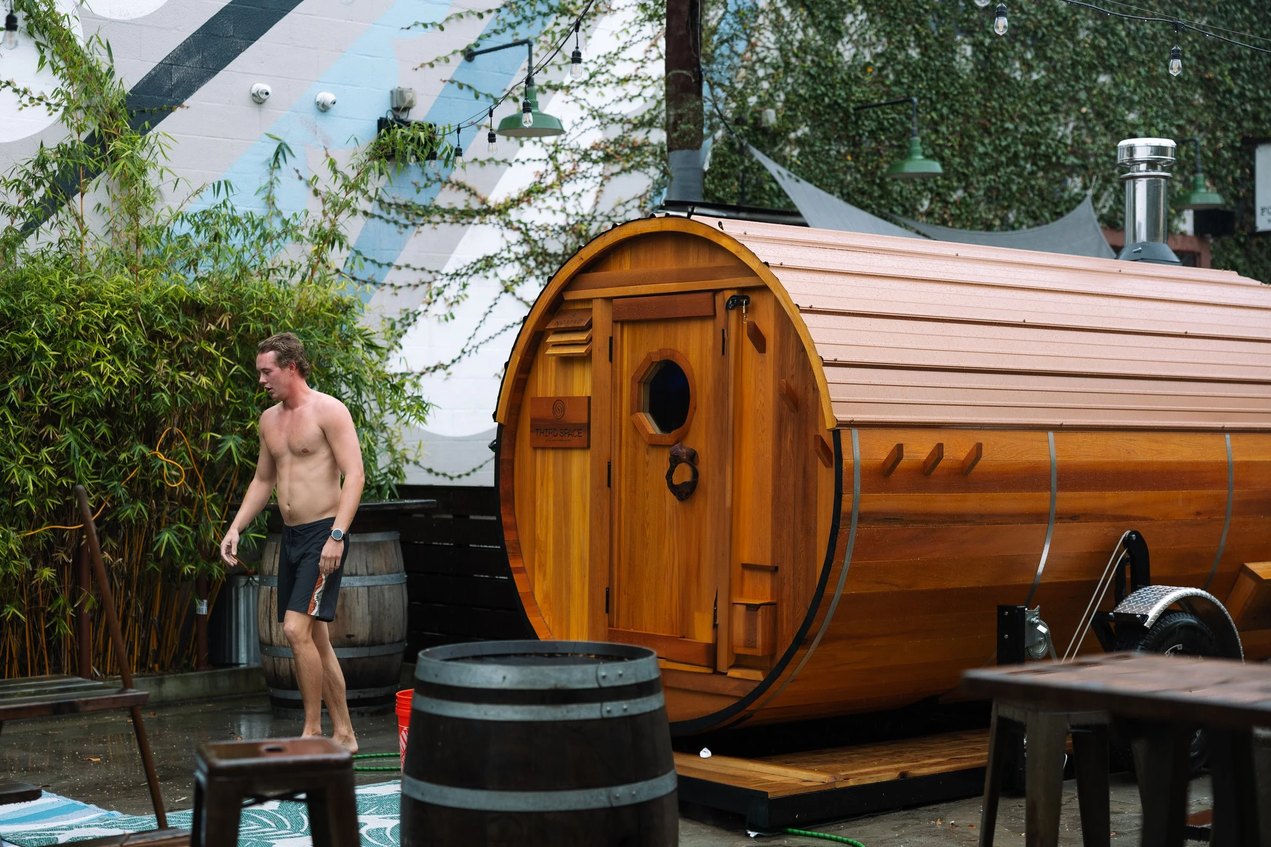 A shirtless man walking past a small wooden sauna trailer in an outdoor setting with bamboo plants and string lights.