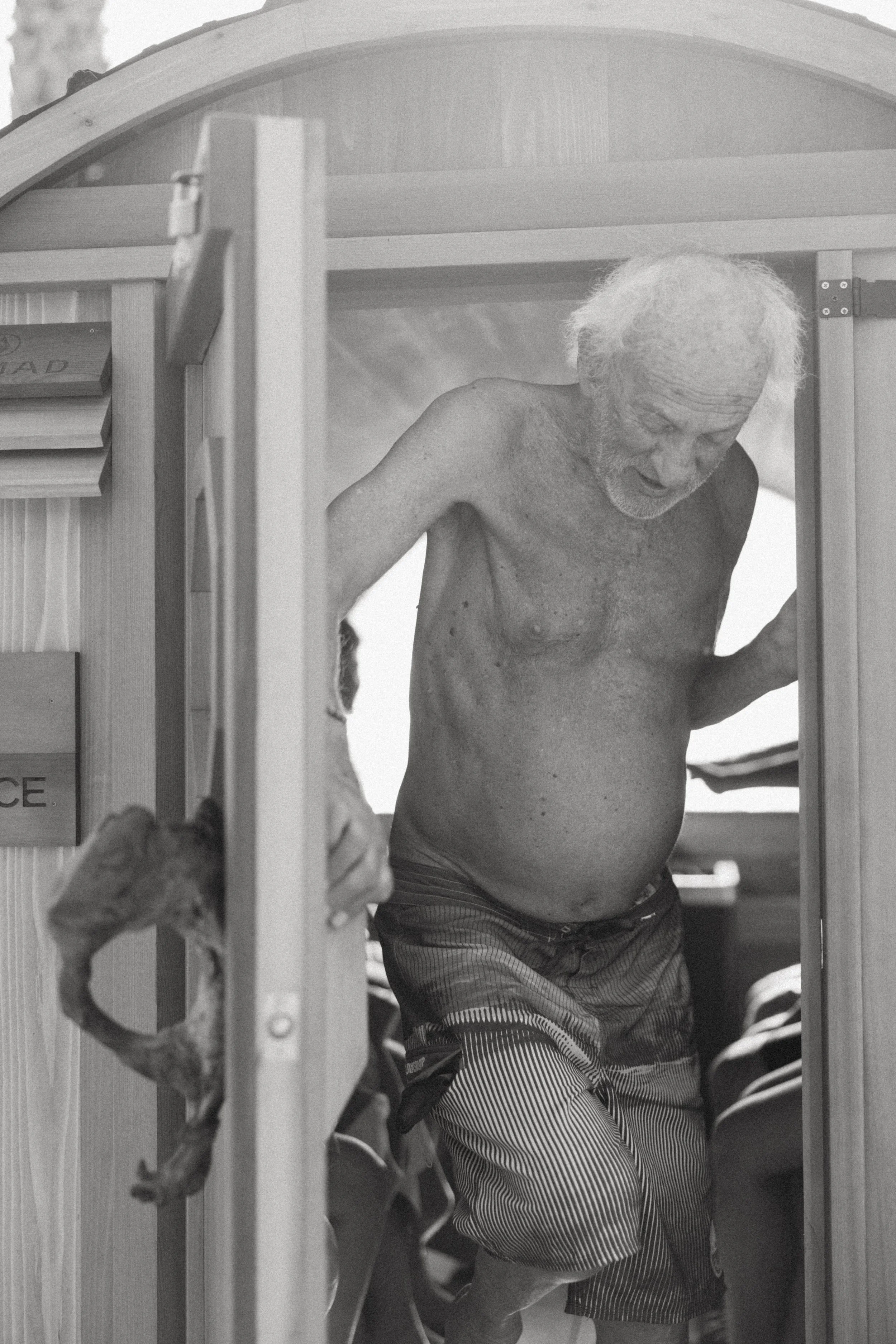 An elderly man with gray hair and a beard, shirtless, is walking through the sauna door in Santa Barbara