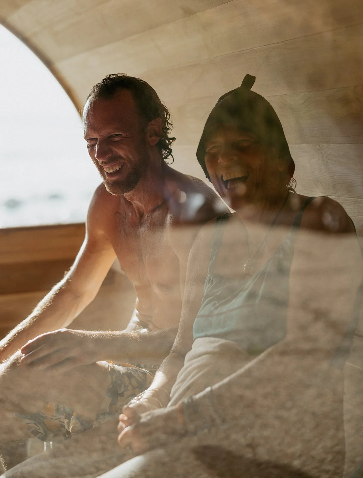 Two people sitting inside a sauna, smiling and laughing, with steam rising around them, and wooden walls in the background.