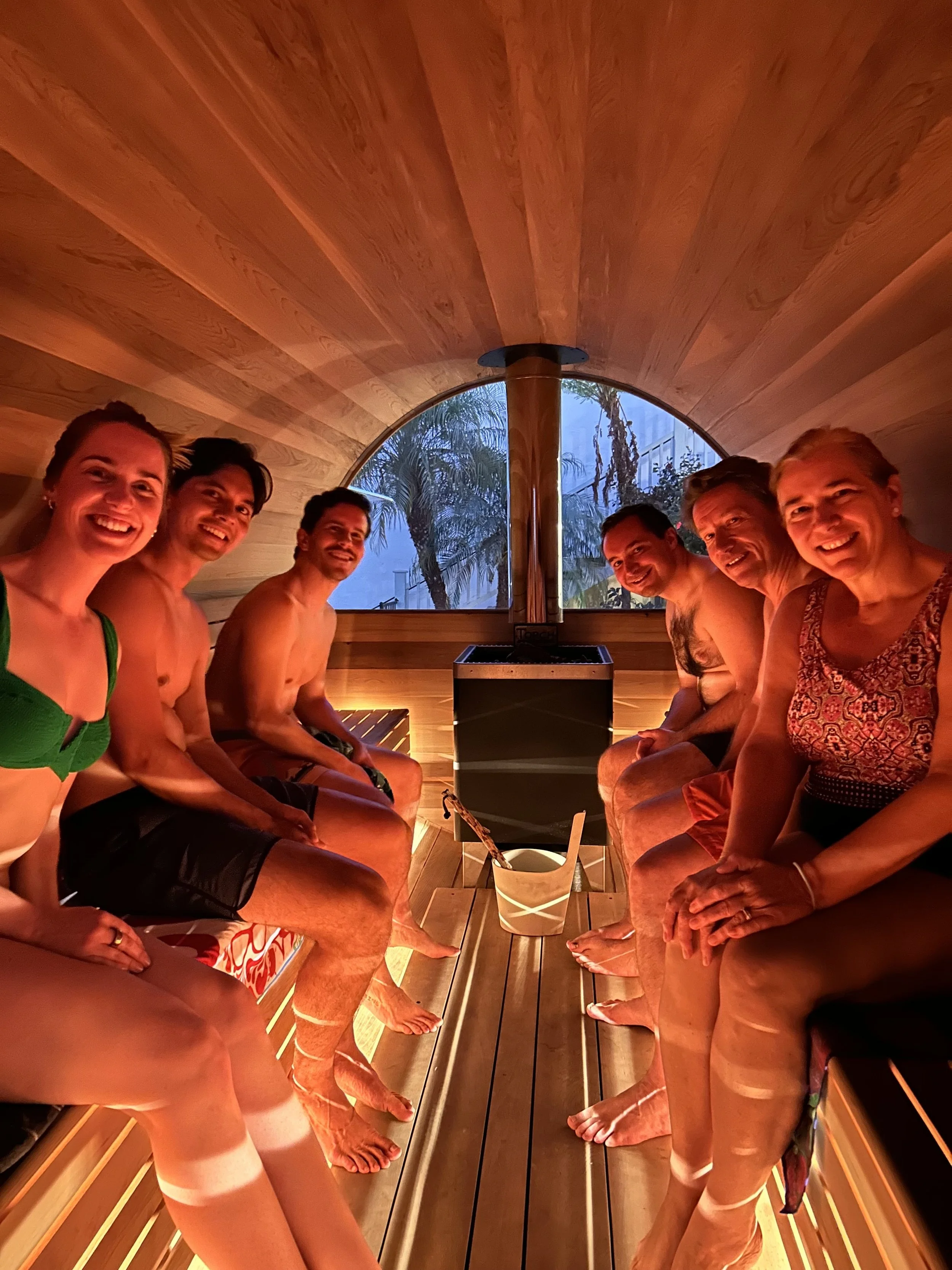 A group of seven people sitting inside a wooden sauna, smiling and looking at the camera, with a small heater in the center and trees visible through a window at the back.