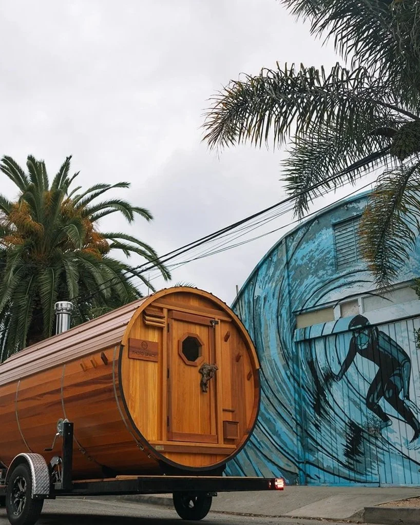 A wooden sauna on a trailer parked in front of a mural depicting a surfer riding a wave.