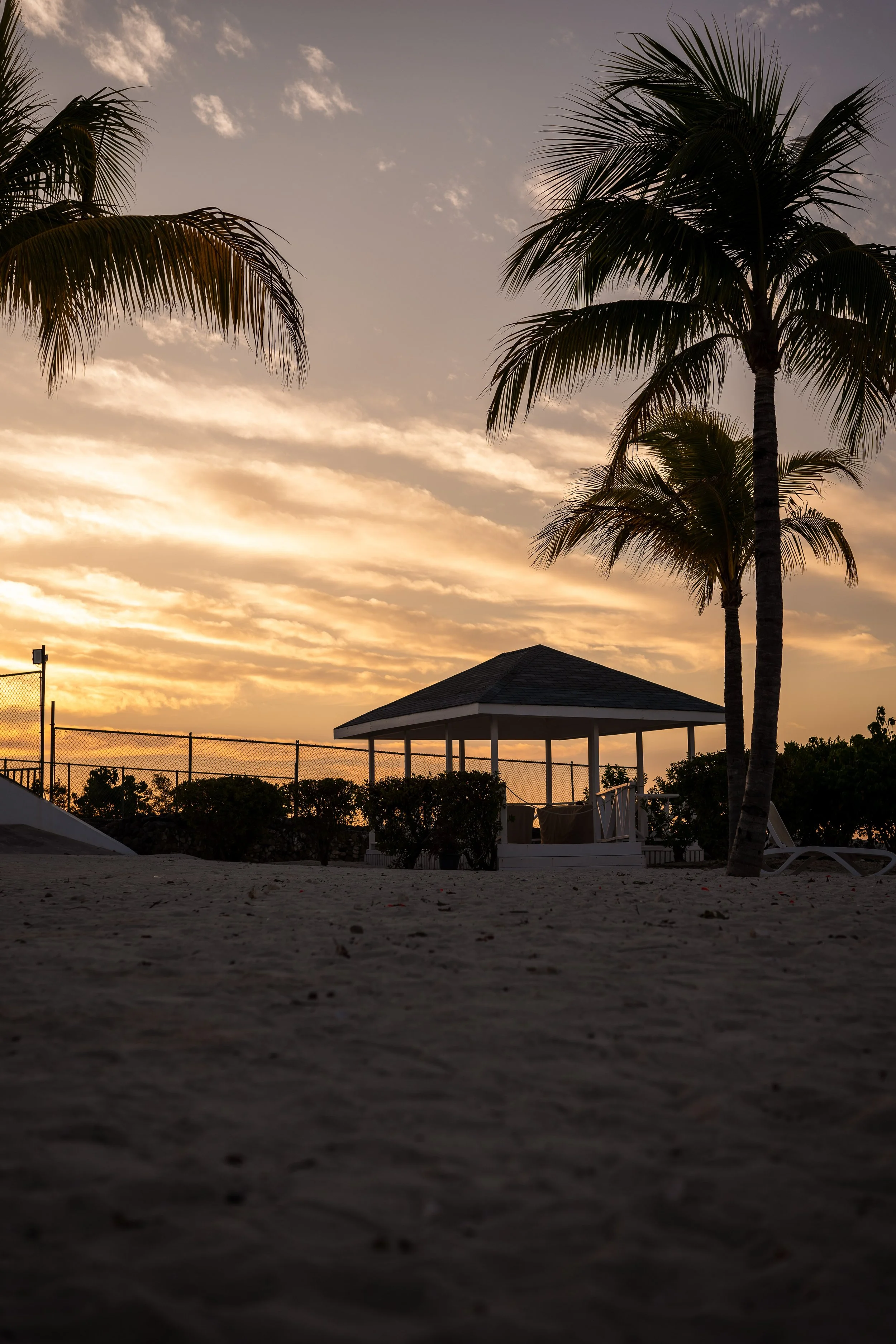 Beach at sunset with palm trees, gazebo, and sandy ground.