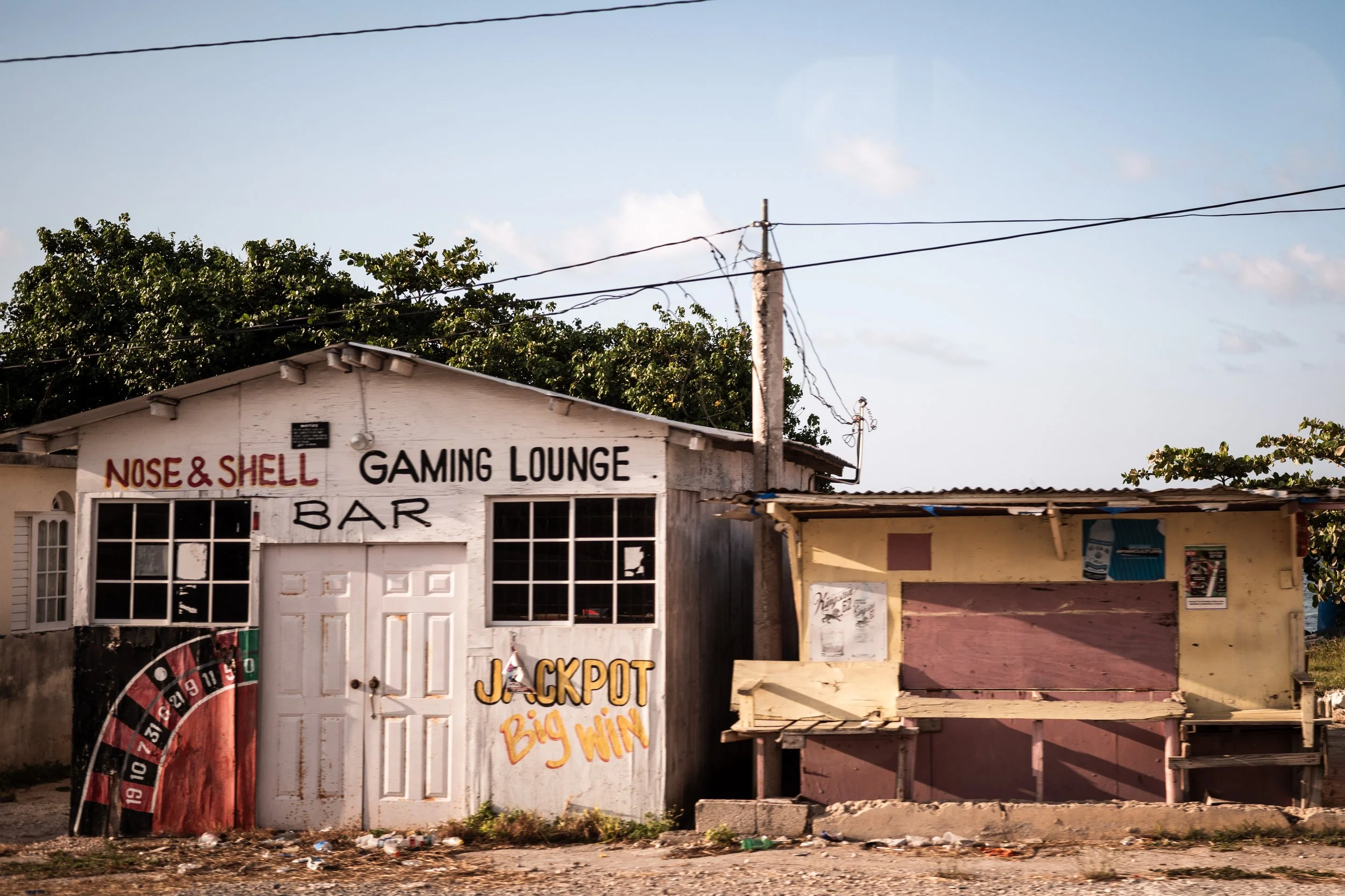 Small building labeled 'Nose & Shell Gaming Lounge Bar' with faded signs and graffiti, located beside a simple wooden structure. Outdoor setting with power lines and trees in the background.