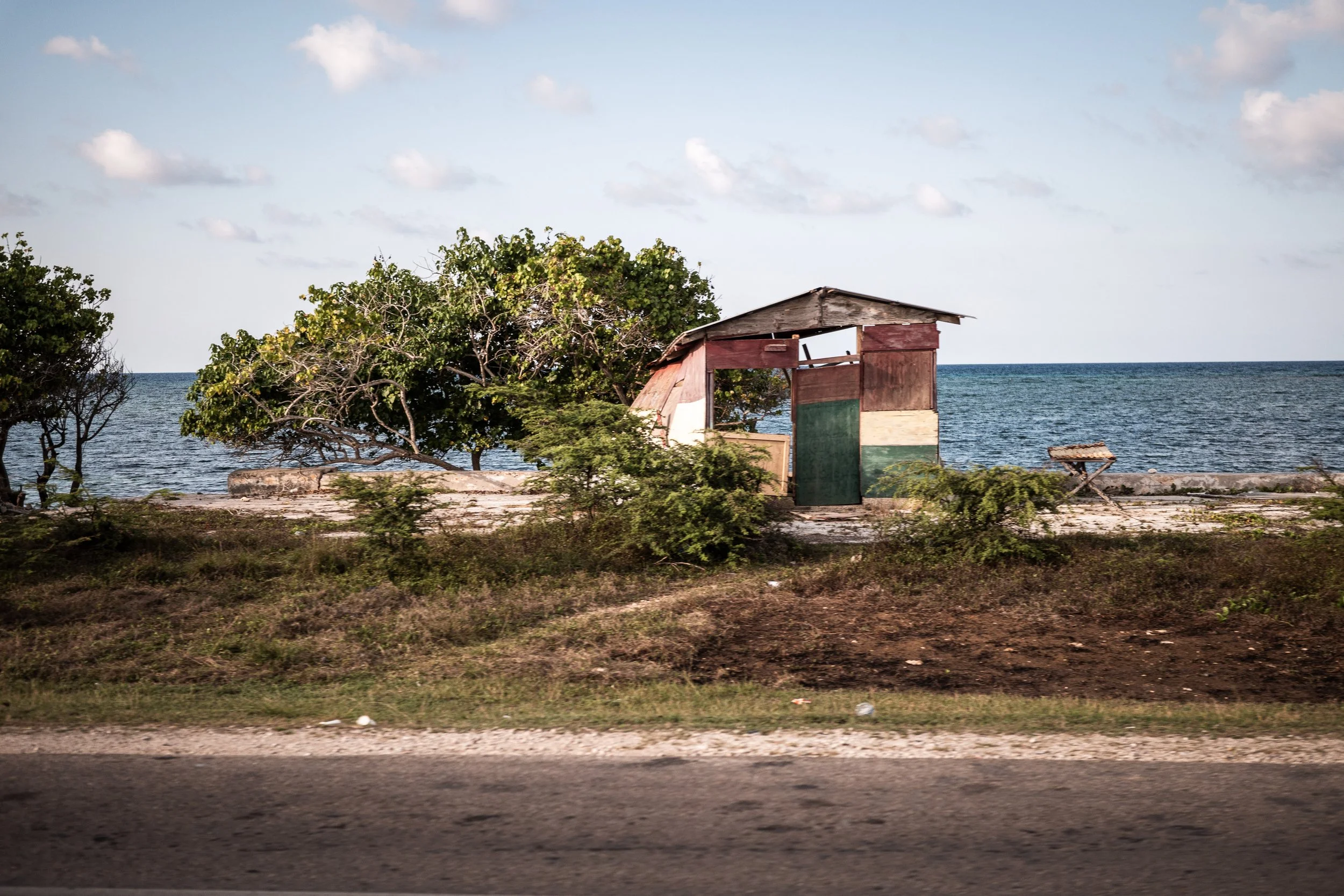 Rustic seaside shack on a beach with ocean in the background, surrounded by trees and sparse vegetation.