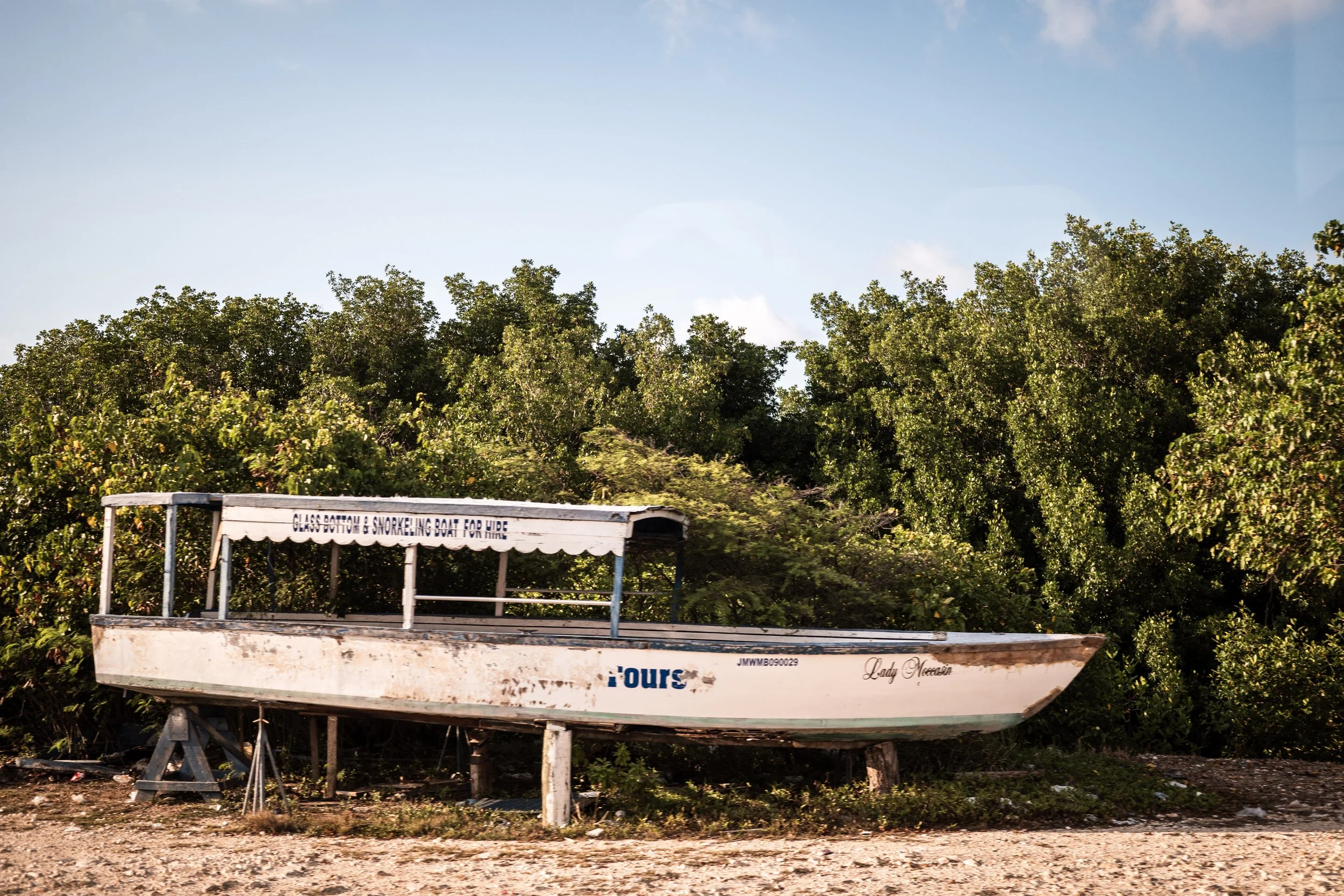 Old white boat named Lady Meow with a canopy, resting on wooden supports on gravel ground, with green bushes in the background and a clear blue sky.