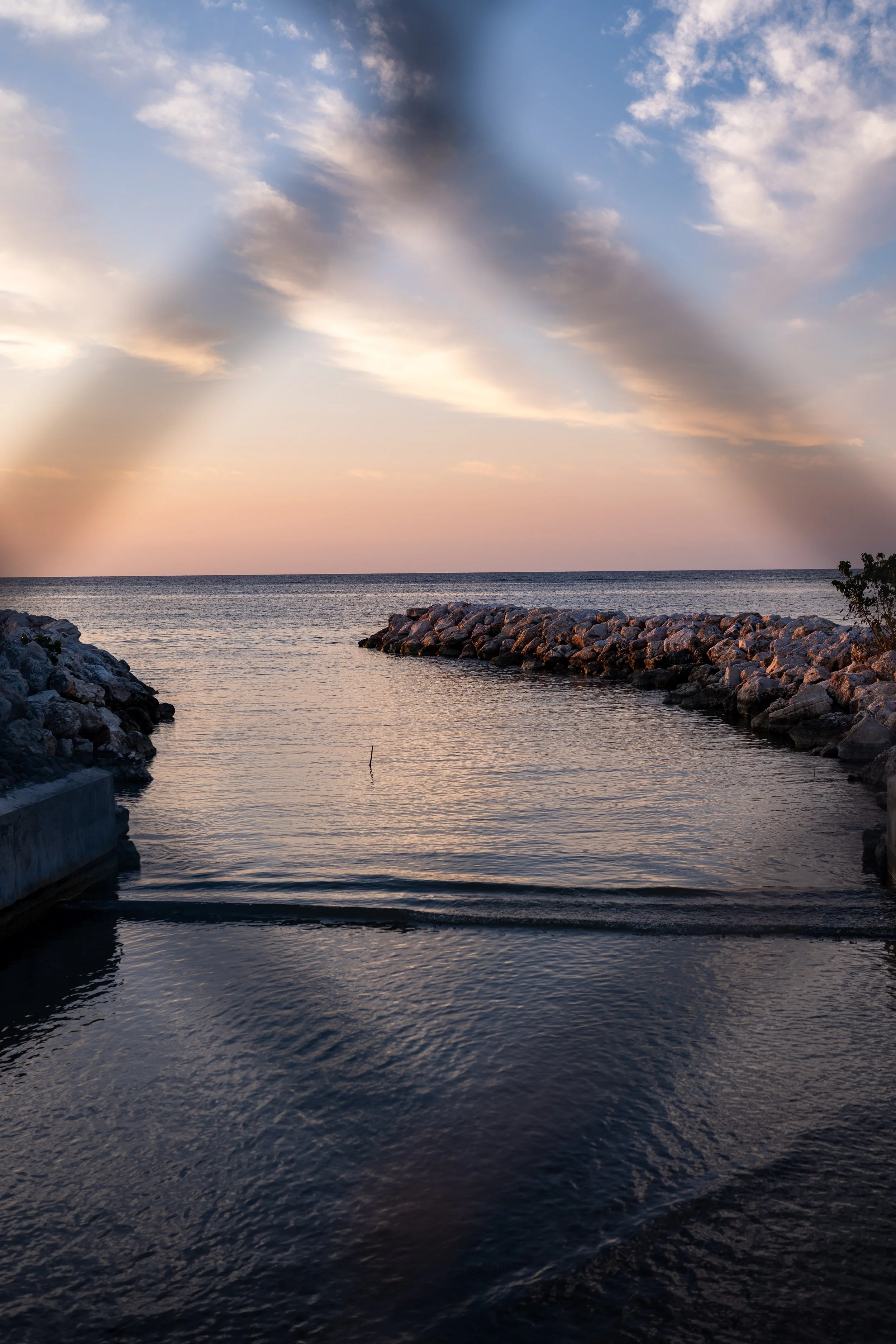 Scenic view of a calm seashore at sunset, with rocks lining the shore and clouds in a colorful sky.