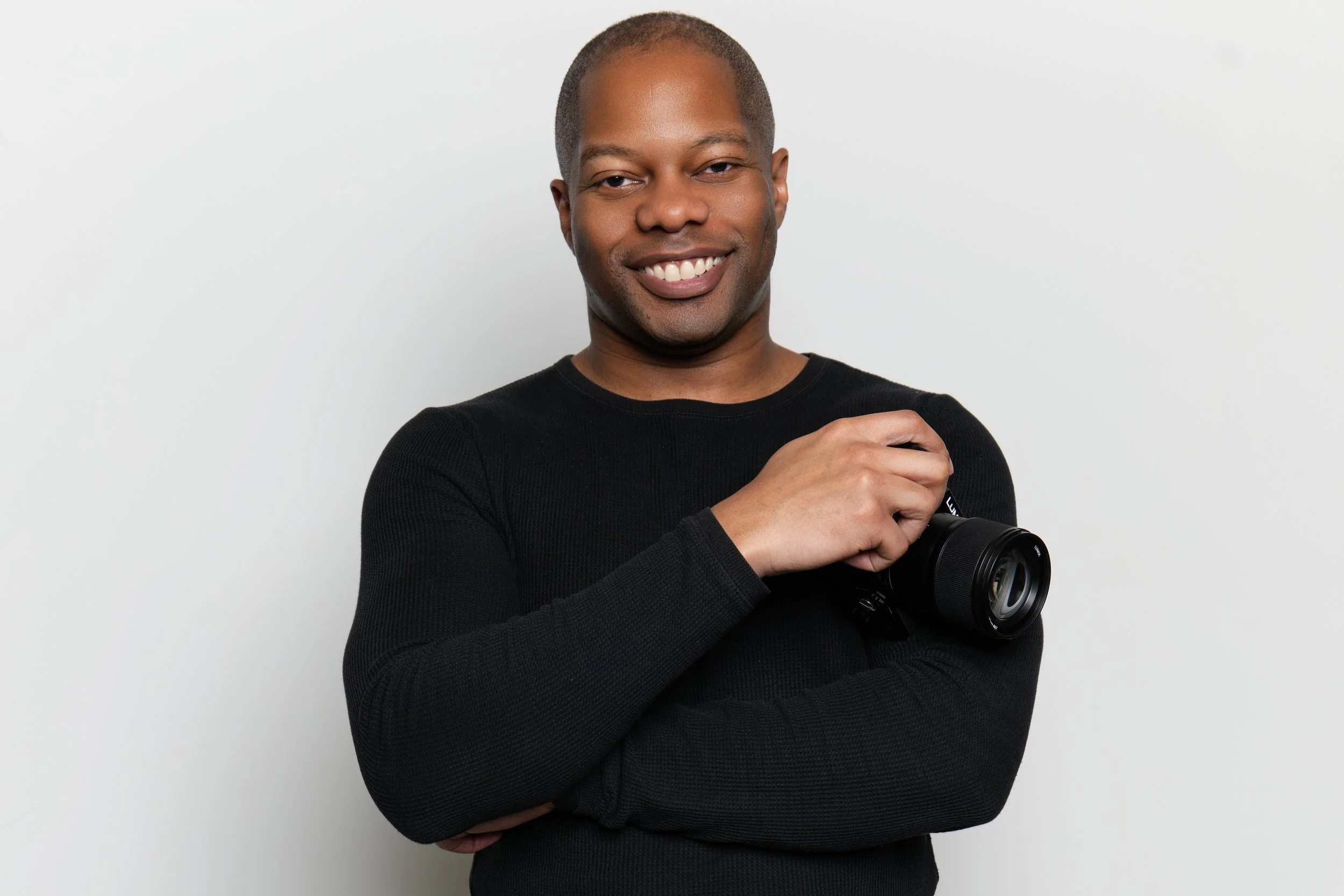 A smiling man in a black long sleeve shirt holding a camera against a plain white background.