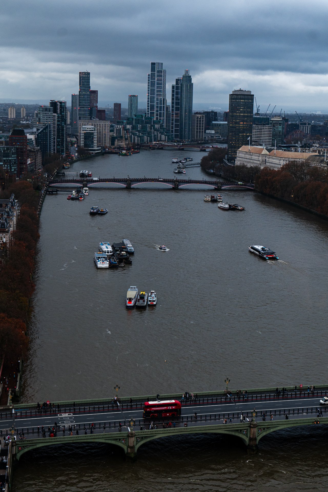 Aerial view of a river in London with multiple bridges, boats, and modern buildings. A red double-decker bus is visible on the nearest bridge.