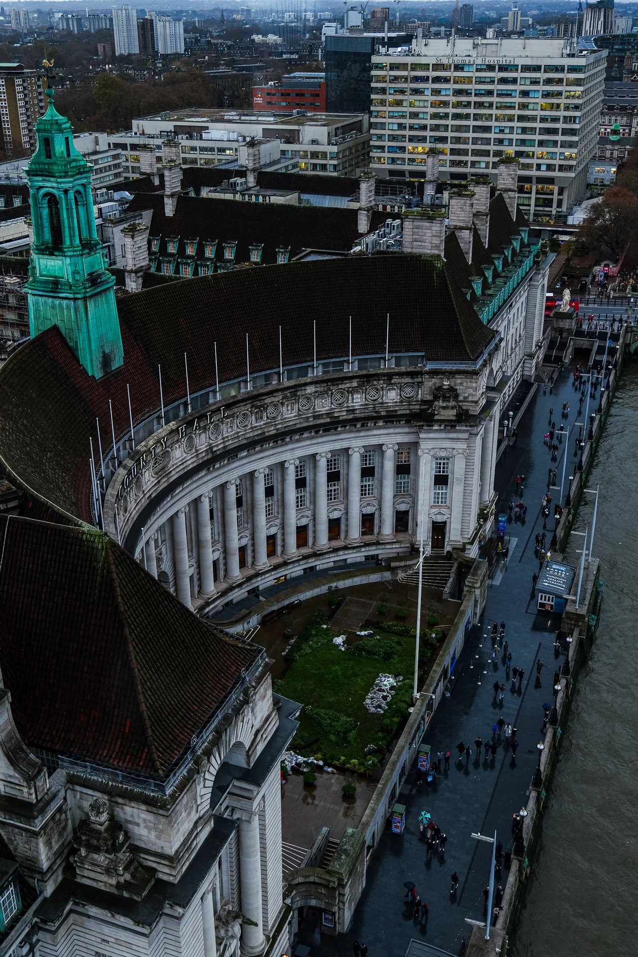 Aerial view of County Hall in London, featuring a curved facade with tall columns and a green copper roof. The surrounding area includes a sidewalk with people walking along the riverbank, tall buildings in the background, and St. Thomas's Hospital i