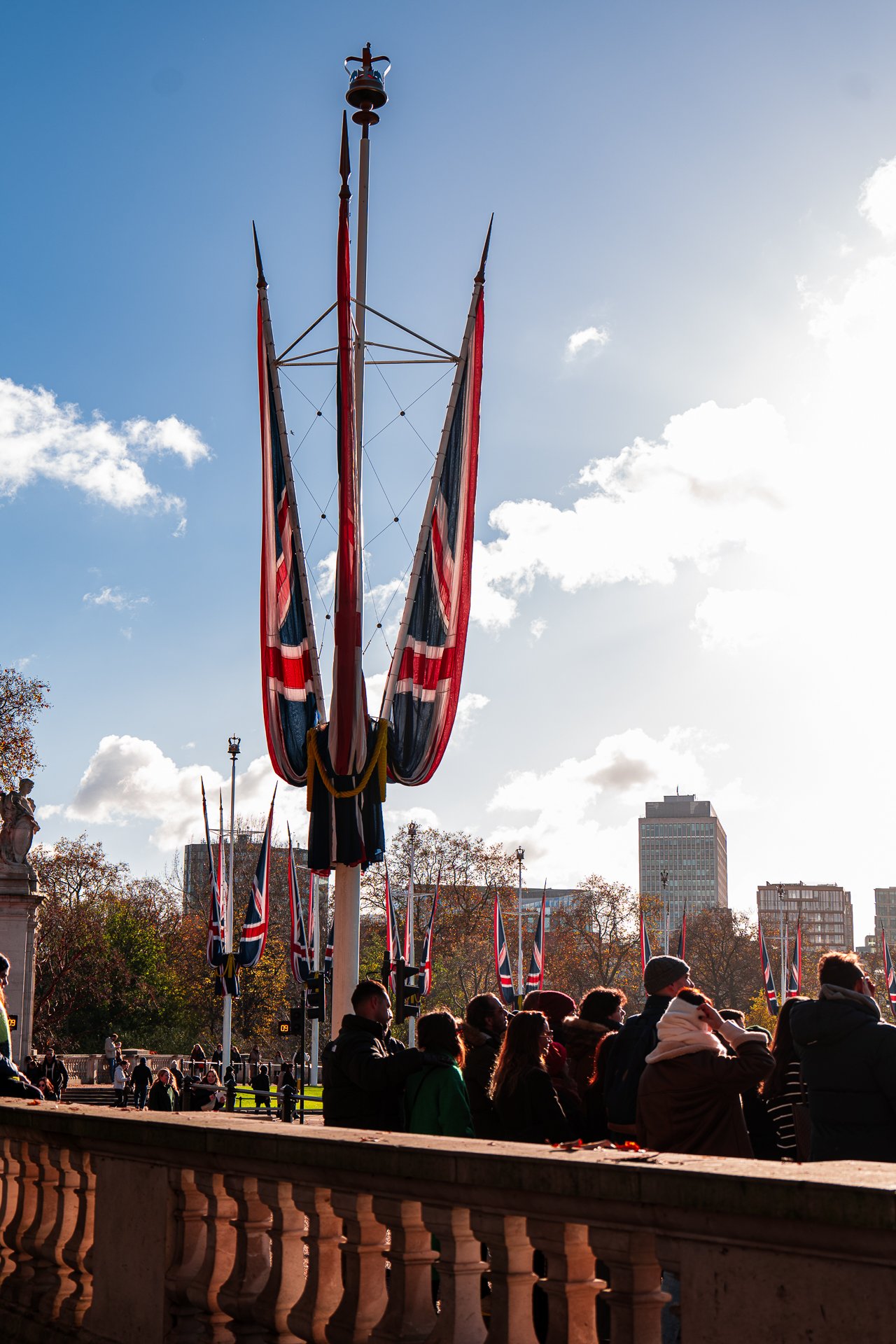 Union Jack flags on poles with crowd of people below, trees and buildings in background, sunny day with few clouds.