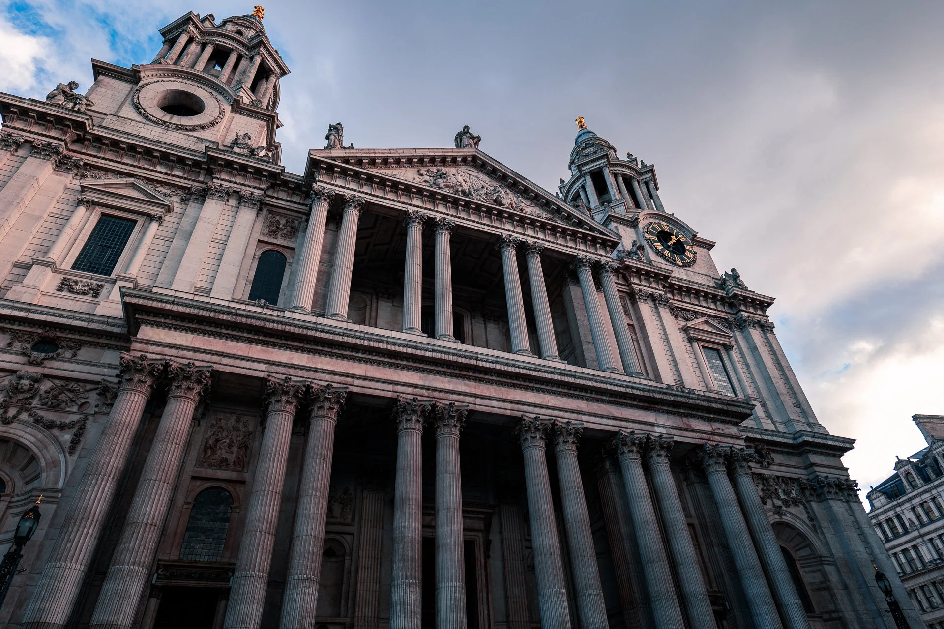 Exterior of St. Paul's Cathedral in London with its iconic columns and clock tower against a cloudy sky.