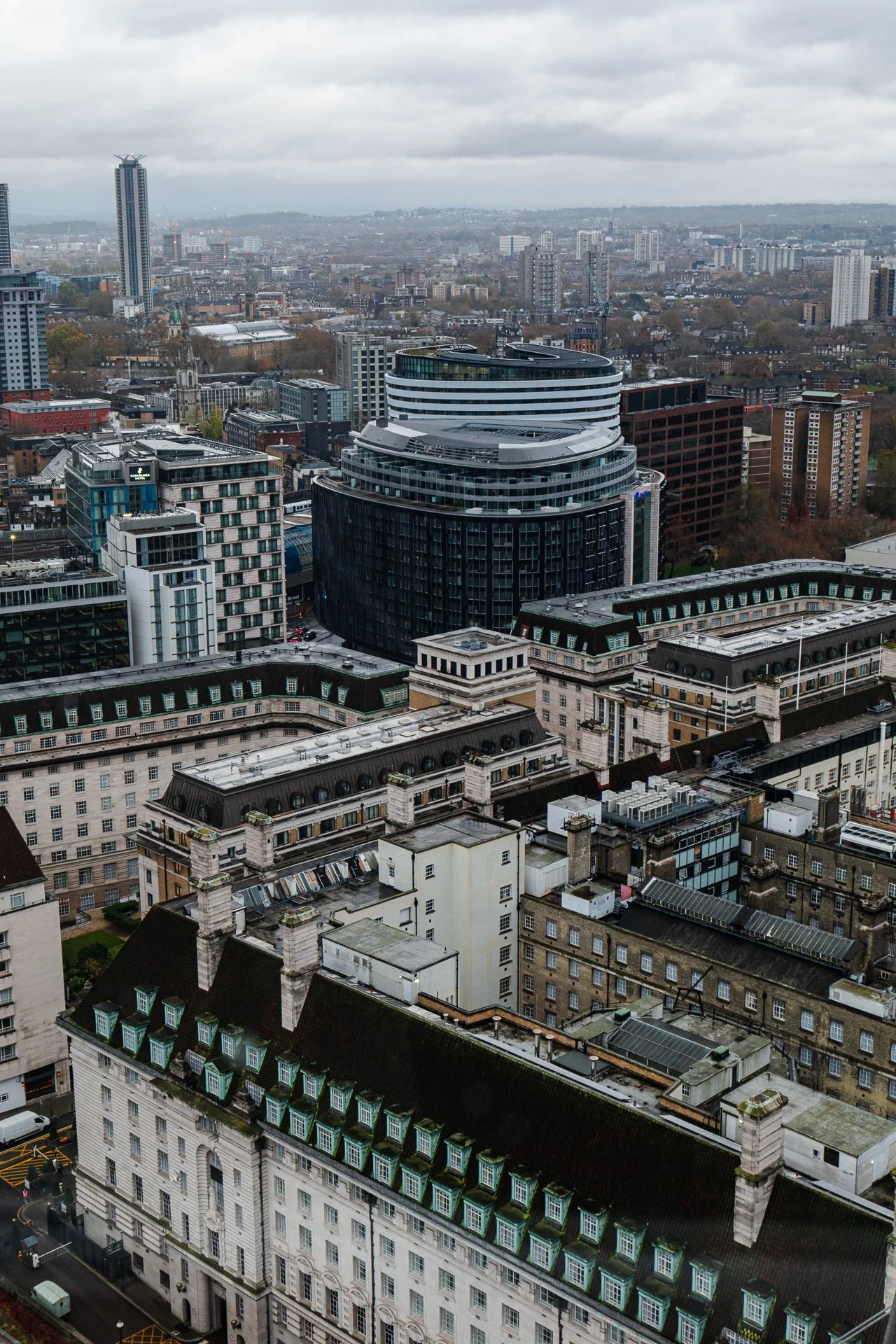 Aerial view of cityscape with modern and classic architecture, featuring a mix of high-rise buildings and traditional-style rooftops, under a cloudy sky.
