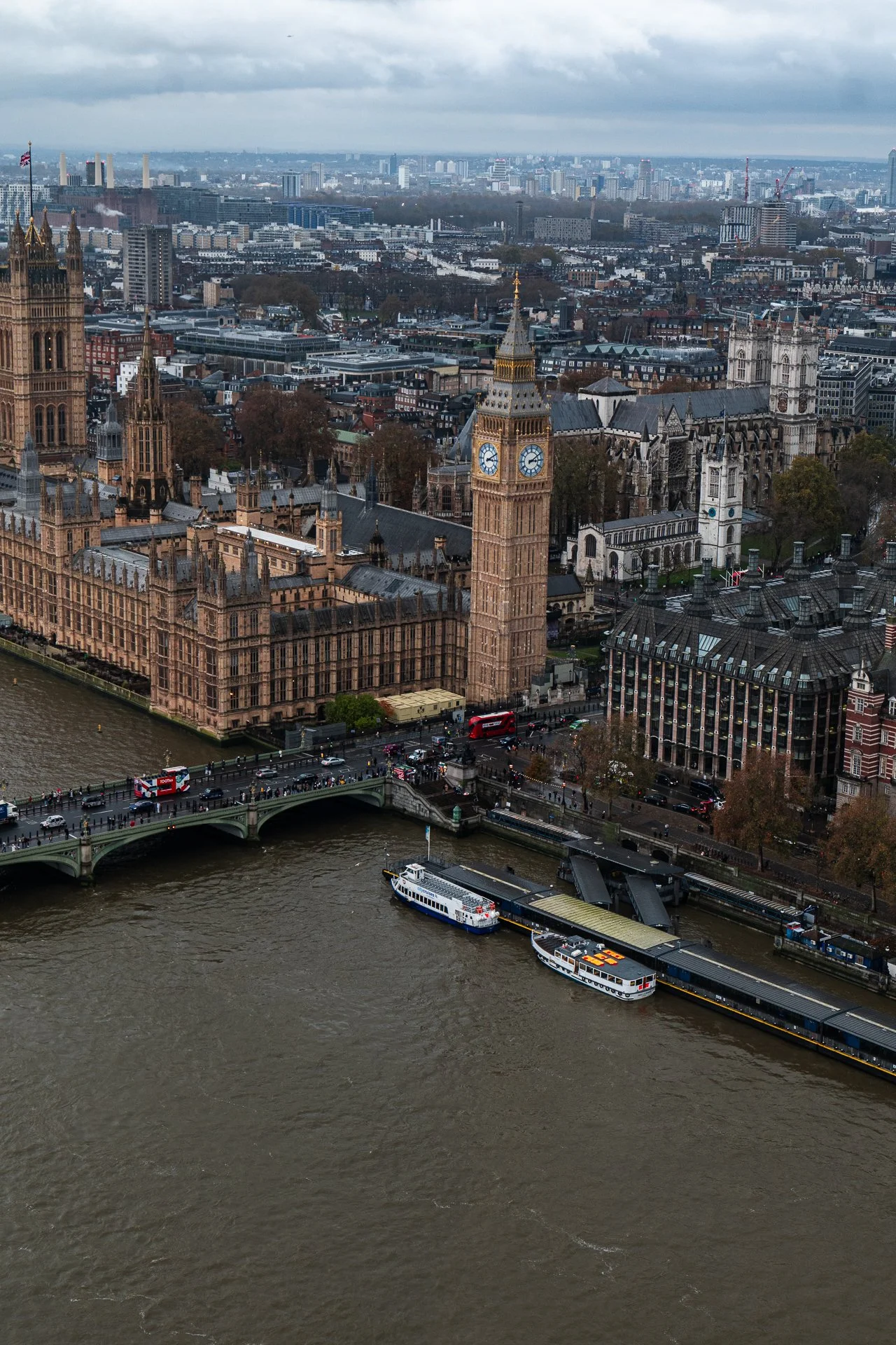 Aerial view of Westminster Palace, Big Ben, and River Thames in London on a cloudy day, with red double-decker buses on Westminster Bridge.