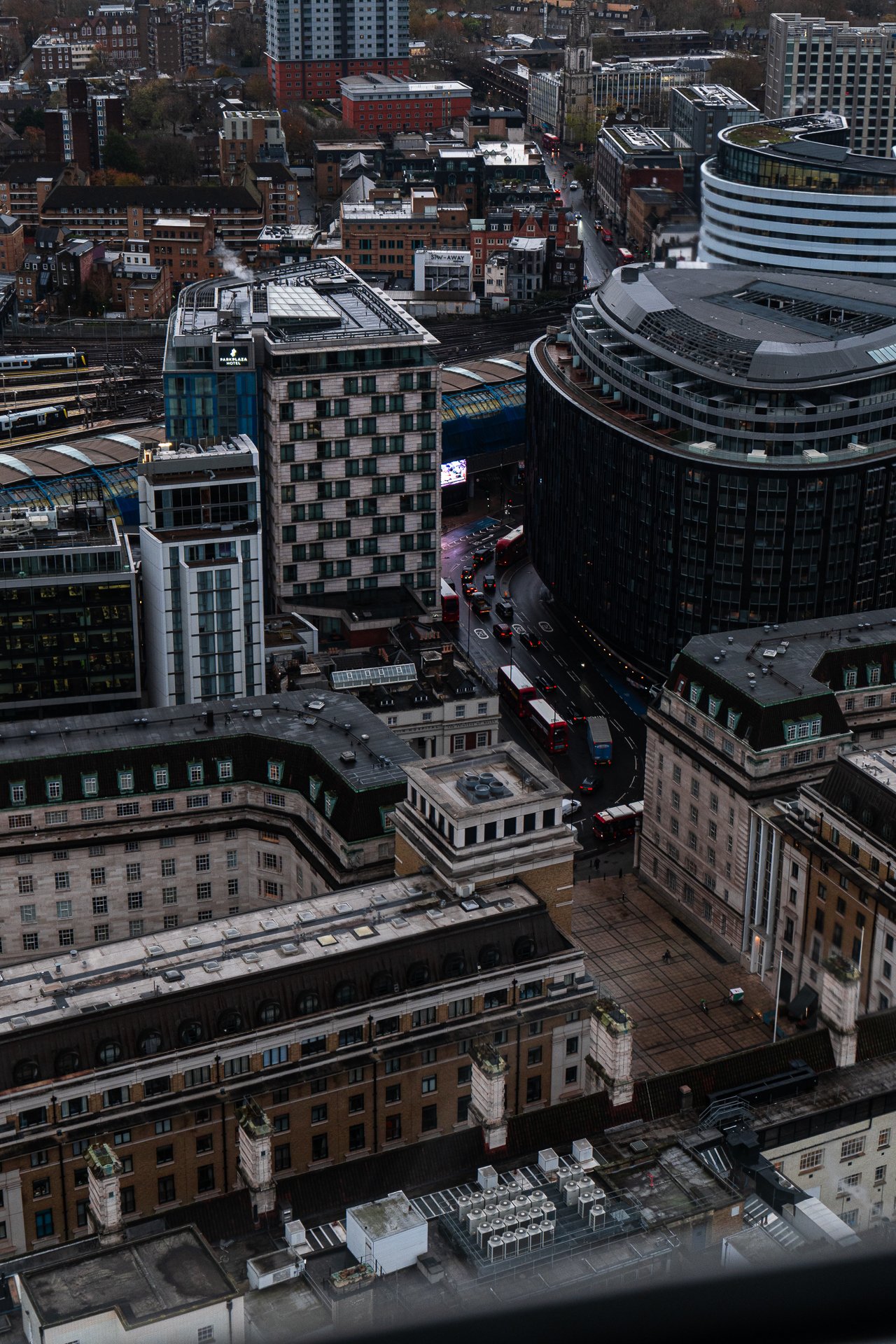 Aerial view of an urban cityscape with modern and classic buildings, a railway station visible, and several red buses in traffic. The mix of architectural styles includes high-rise structures and traditional brick buildings. A network of roads and tr