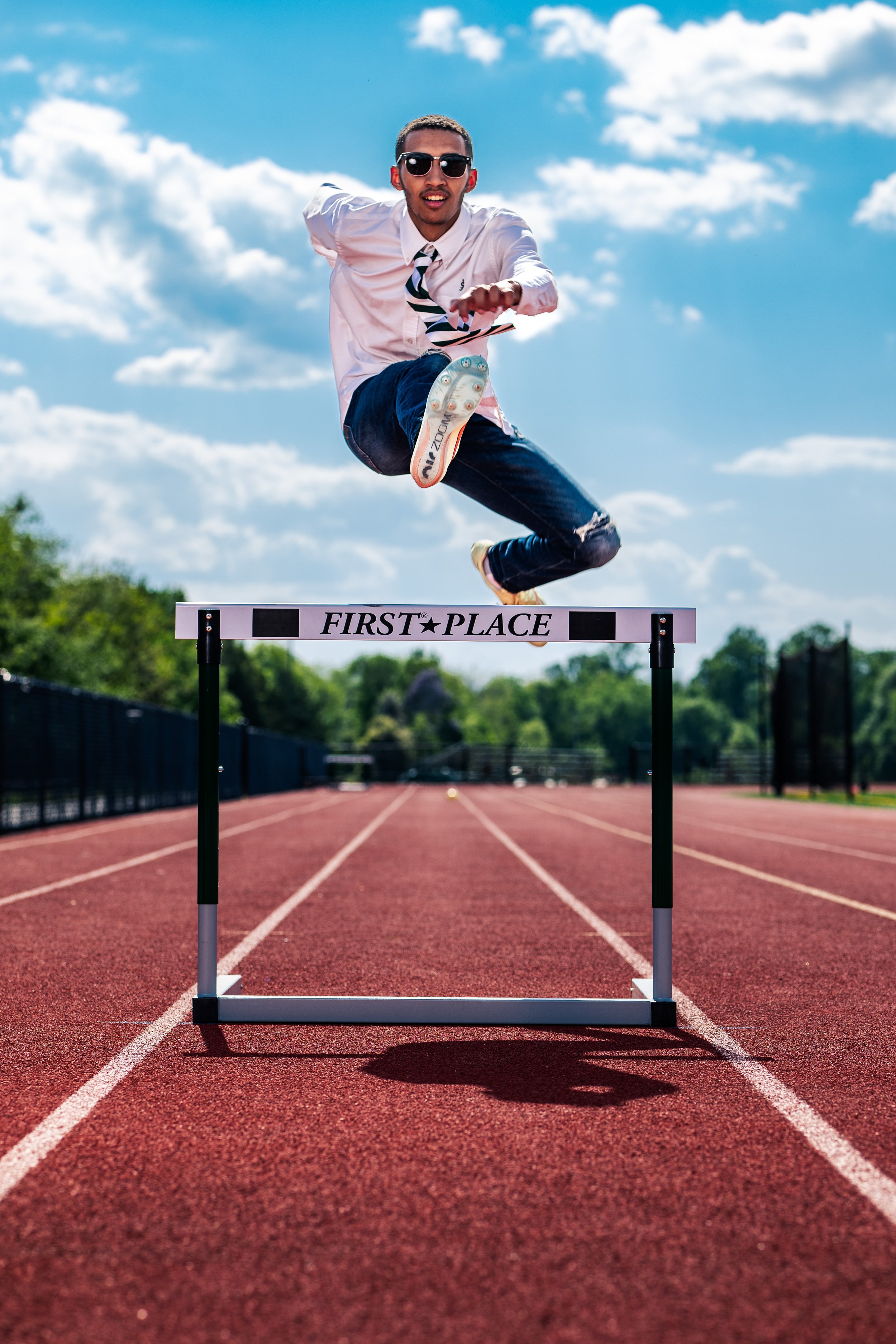 A man jumping over a hurdle on an outdoor running track, wearing sunglasses, a white shirt, a striped tie, and jeans, with a blue sky and scattered clouds in the background.