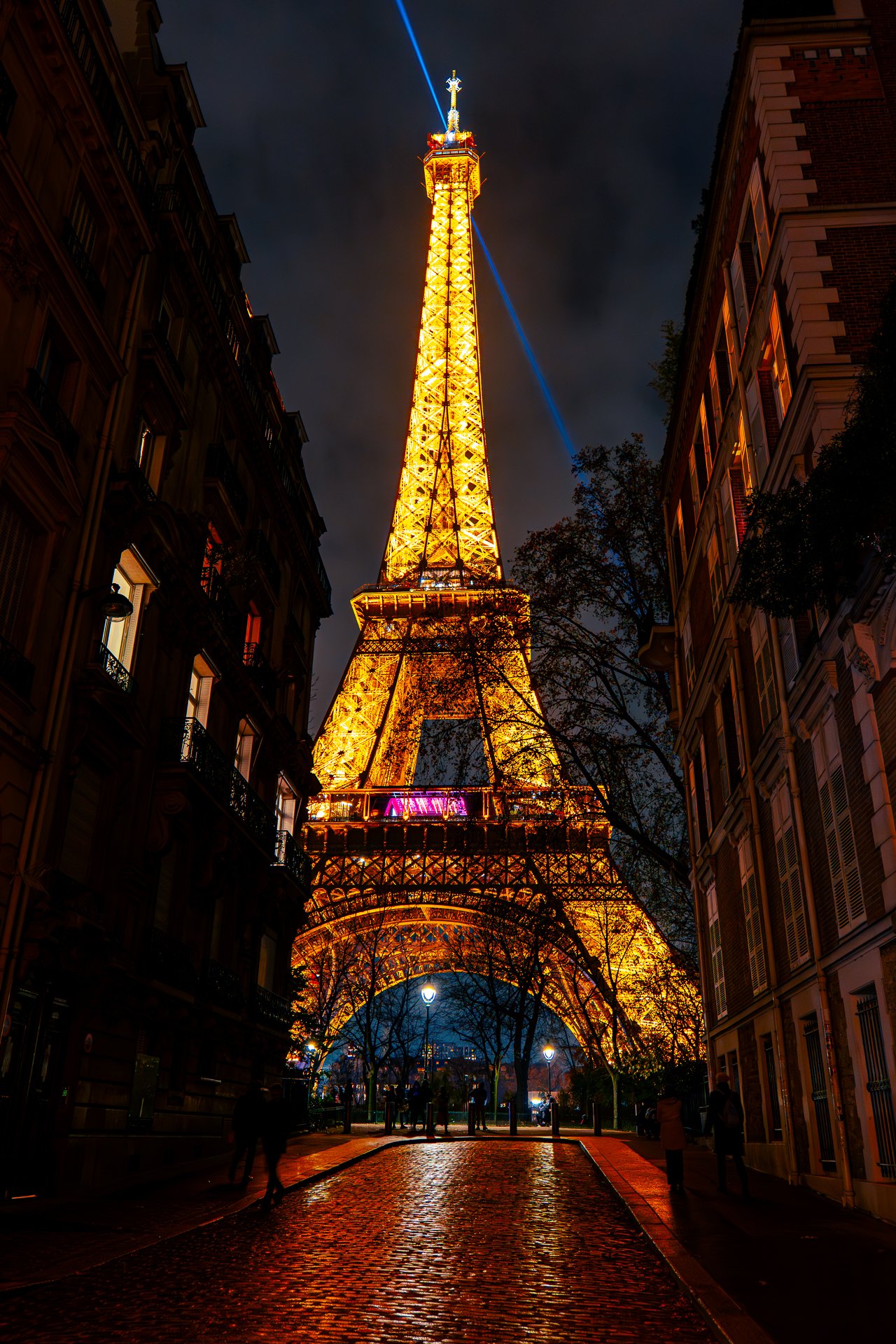 Eiffel Tower illuminated at night, viewed from a cobblestone street between two buildings, with a blue spotlight at the top.