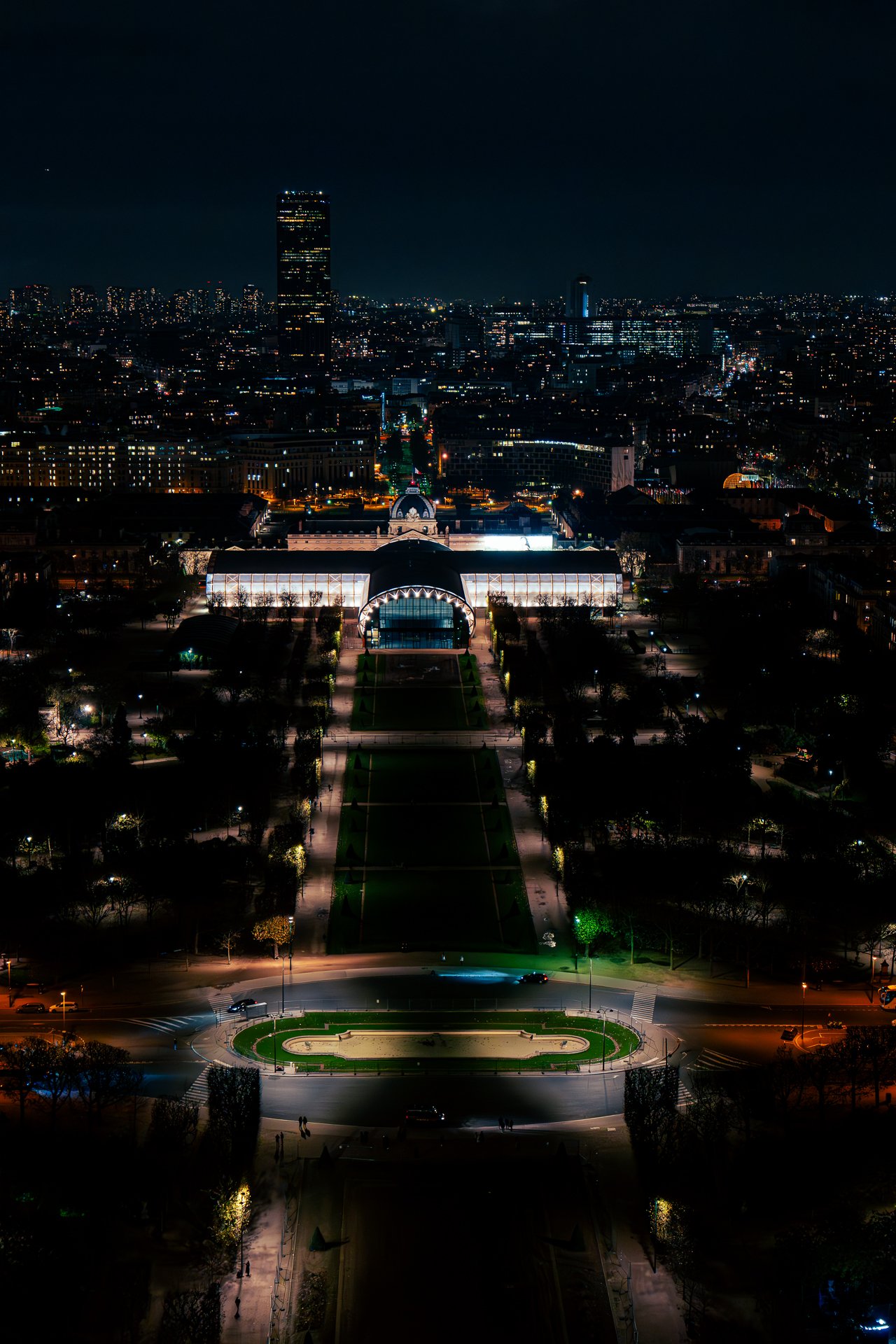 Aerial view of a cityscape at night, featuring a well-lit, symmetrical garden leading to a large building with a domed roof. Skyscrapers and city lights are visible in the background.
