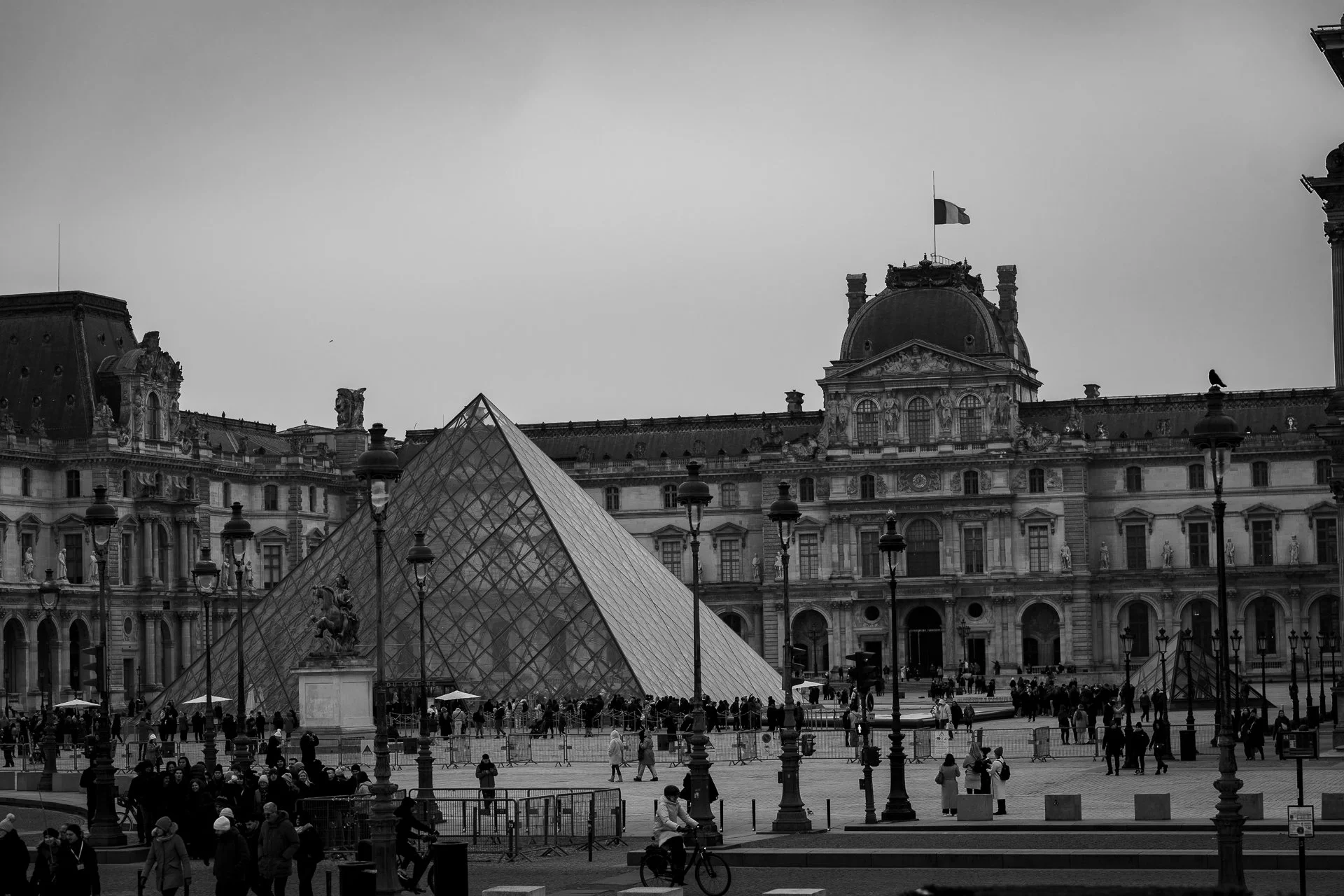 Black and white photo of the Louvre Museum and glass pyramid entrance in Paris, with people walking in the square.