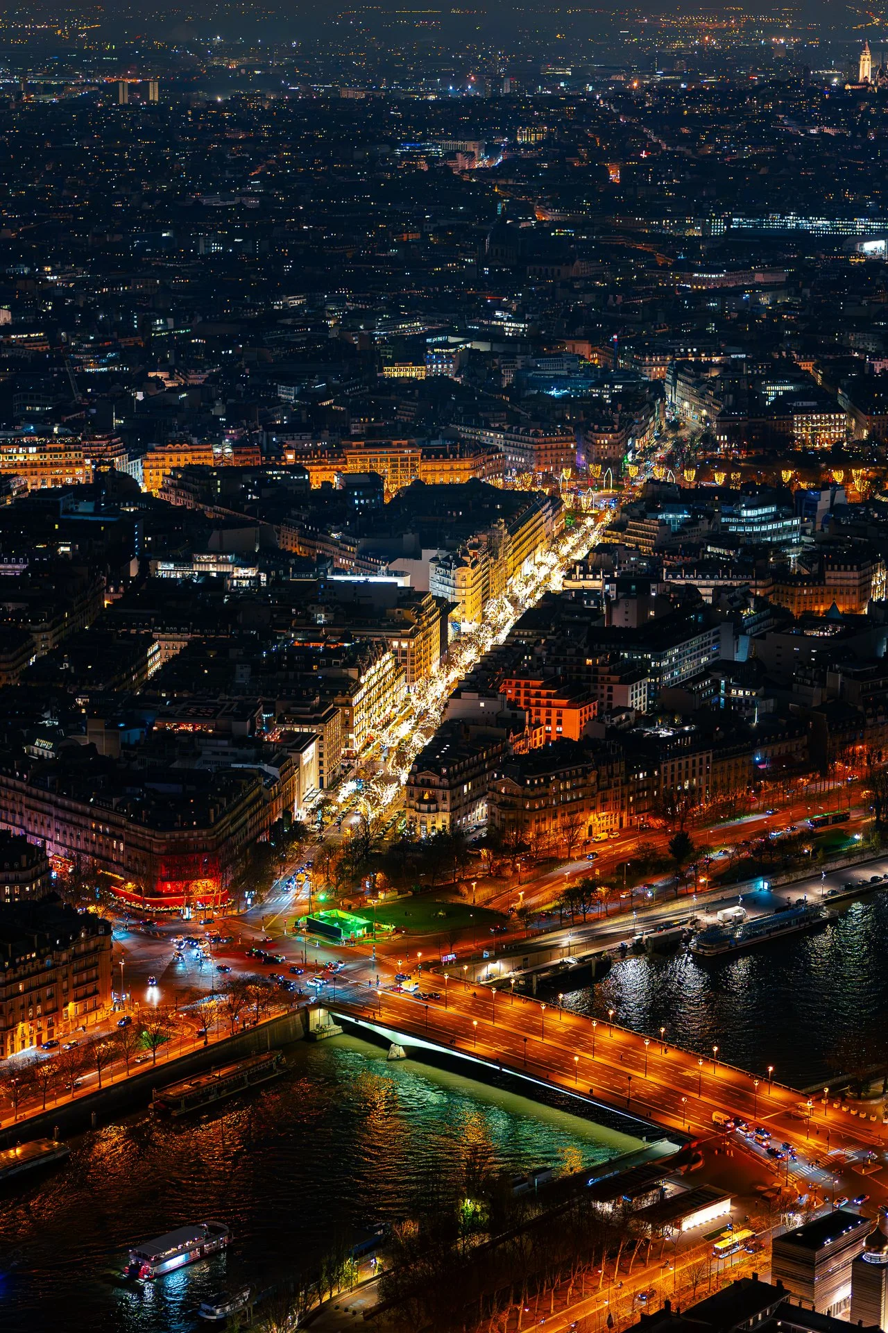 Aerial view of a cityscape at night with illuminated streets and buildings, bridge crossing a river, and bustling traffic.