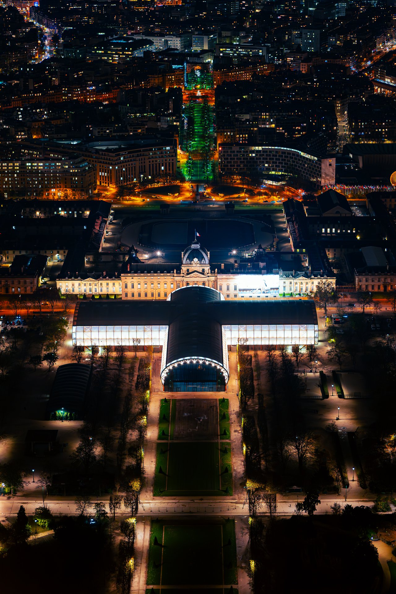 Aerial night view of a cityscape featuring a brightly illuminated building with a distinctive glass roof, surrounded by landscaped gardens and tree-lined paths. The city lights create a glowing backdrop in the distance.