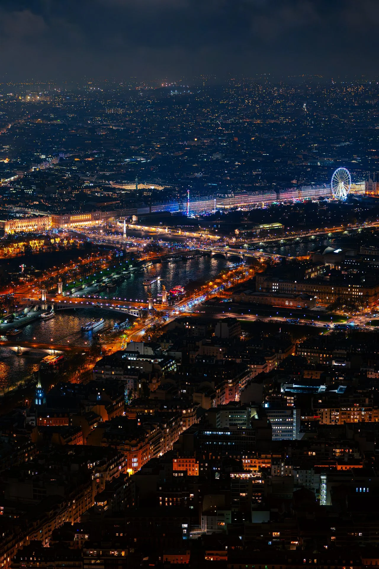 Aerial night view of Paris with illuminated streets, bridges, and a large Ferris wheel.