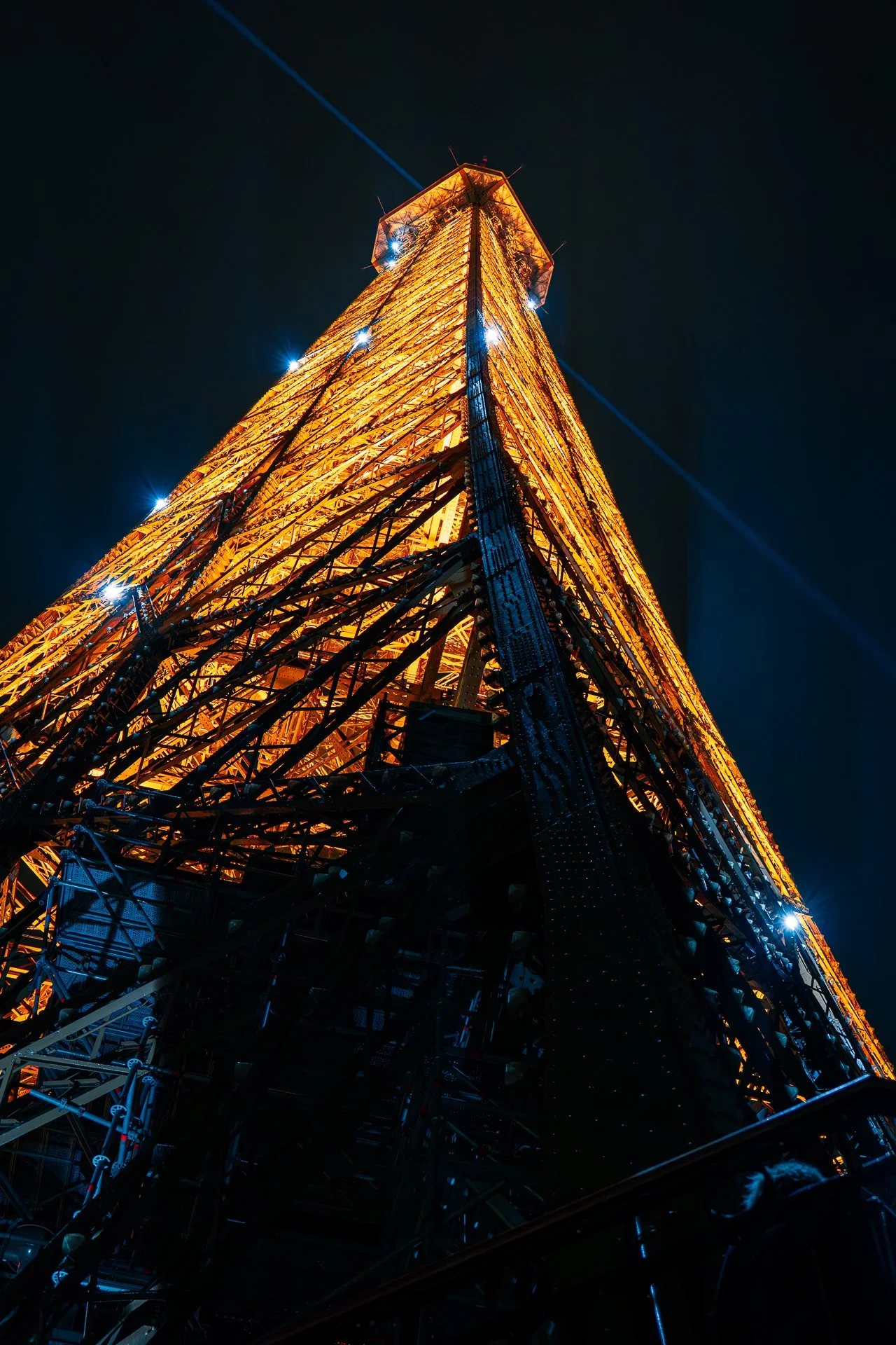 View of a tall illuminated metal tower at night, looking upwards from the base.