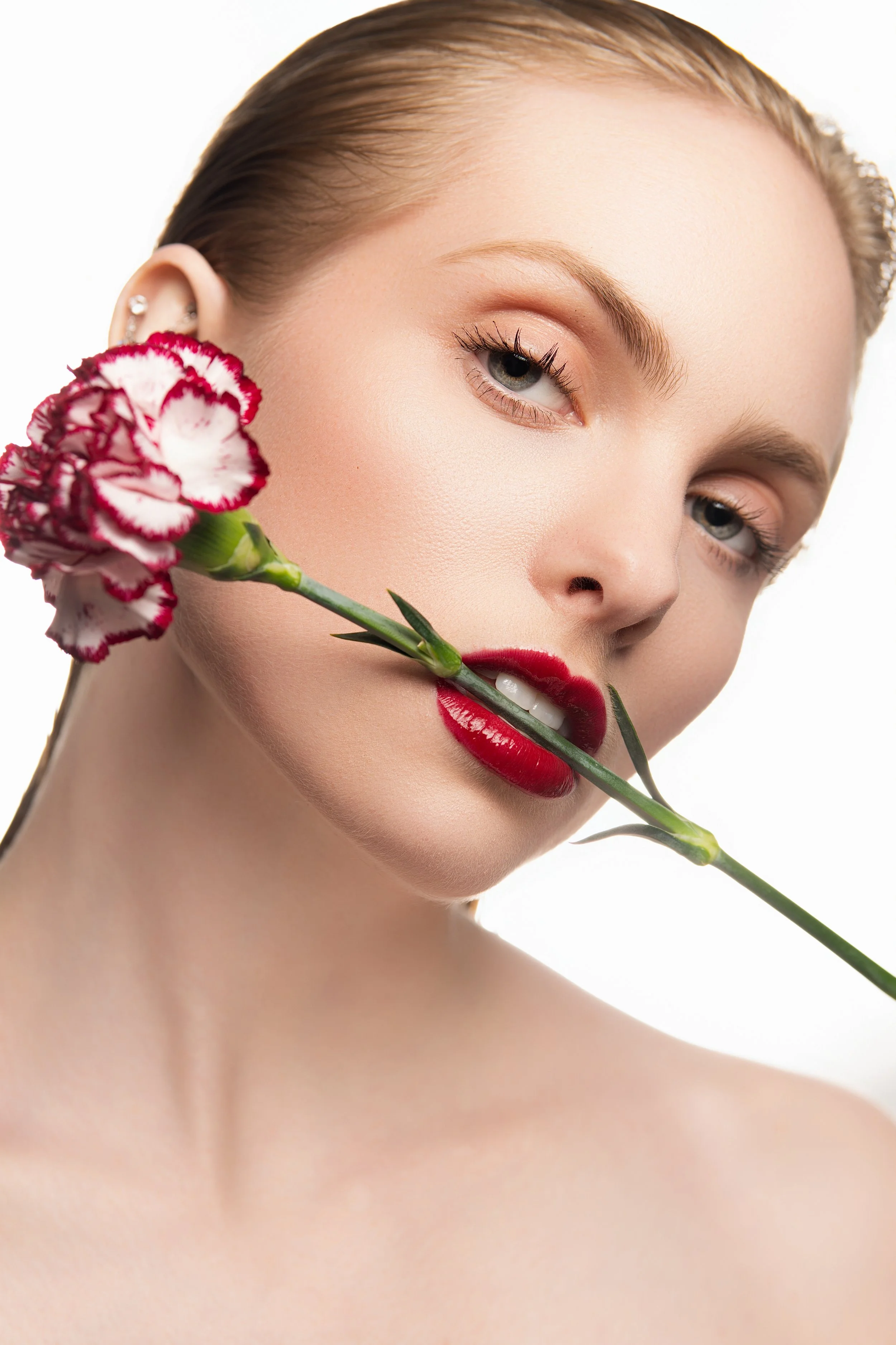 A woman with blonde hair and red lipstick holding a pink and white carnation flower near her face, with a neutral expression against a white background.
