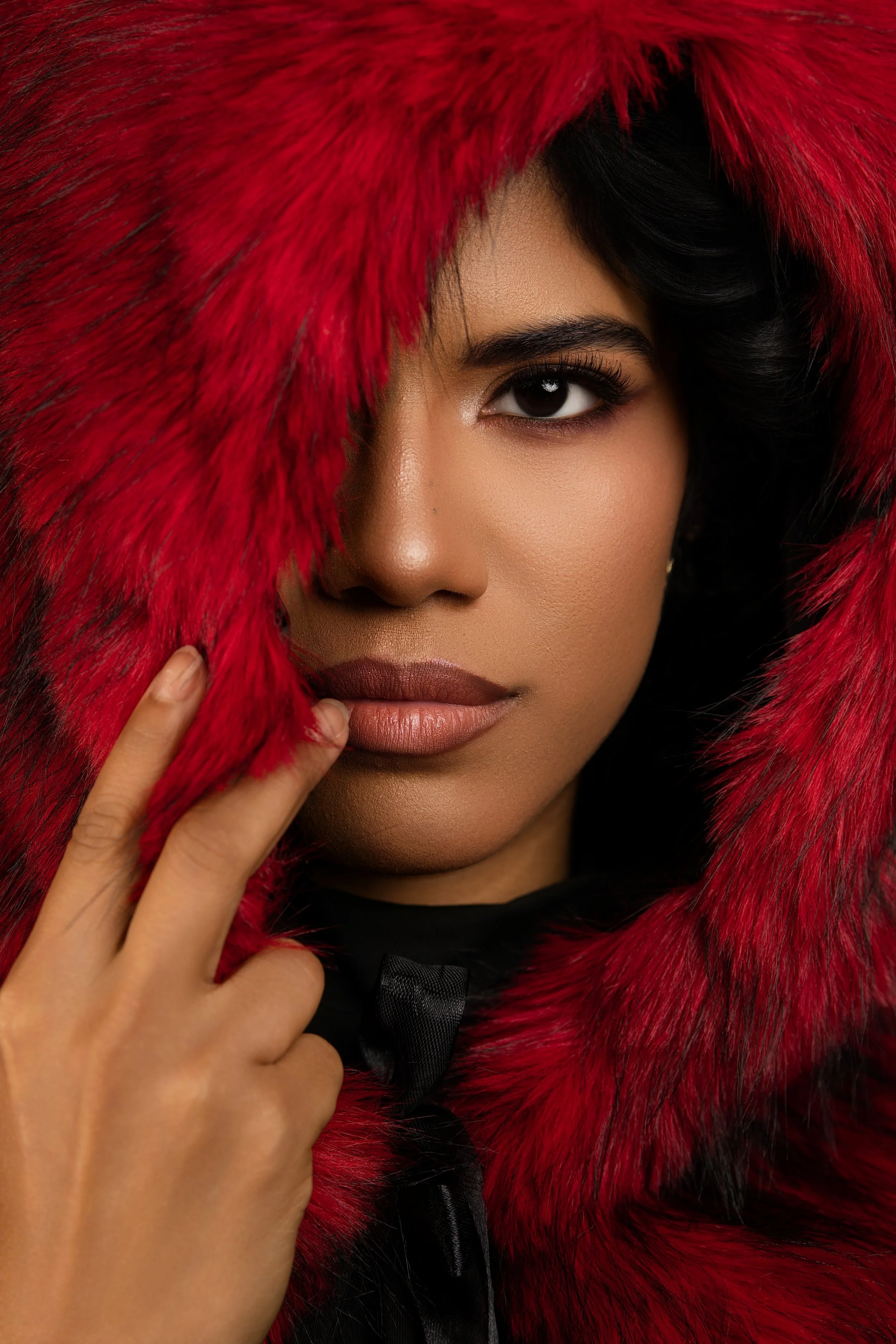 A close-up of a woman's face partially covered by a red fur hood, with dark hair and makeup highlighting her eye and lips.