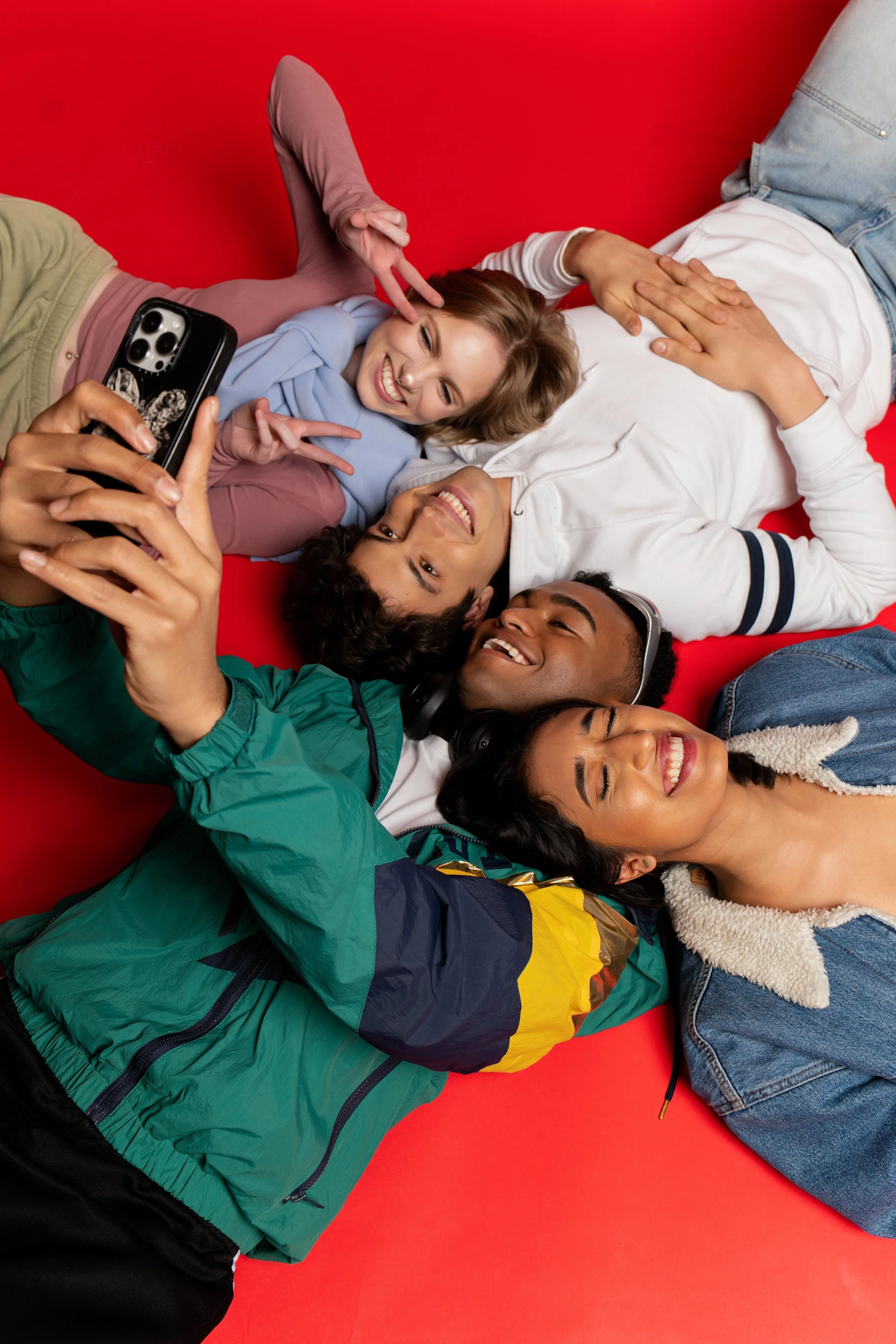 Group of friends lying on red background, taking a selfie, smiling, wearing casual clothes.