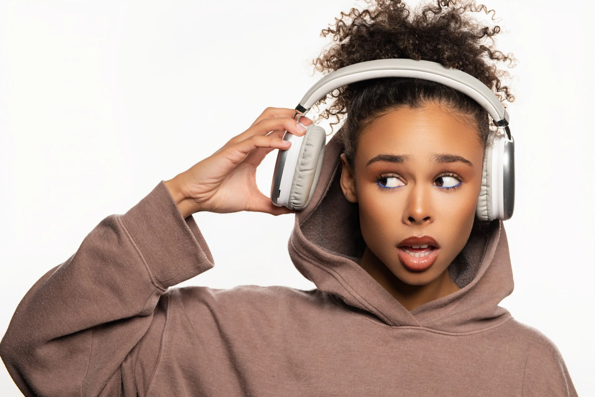 A young woman with curly hair wearing a brown hoodie and listening to music on large over-ear headphones, looking slightly to the side with her mouth slightly open.