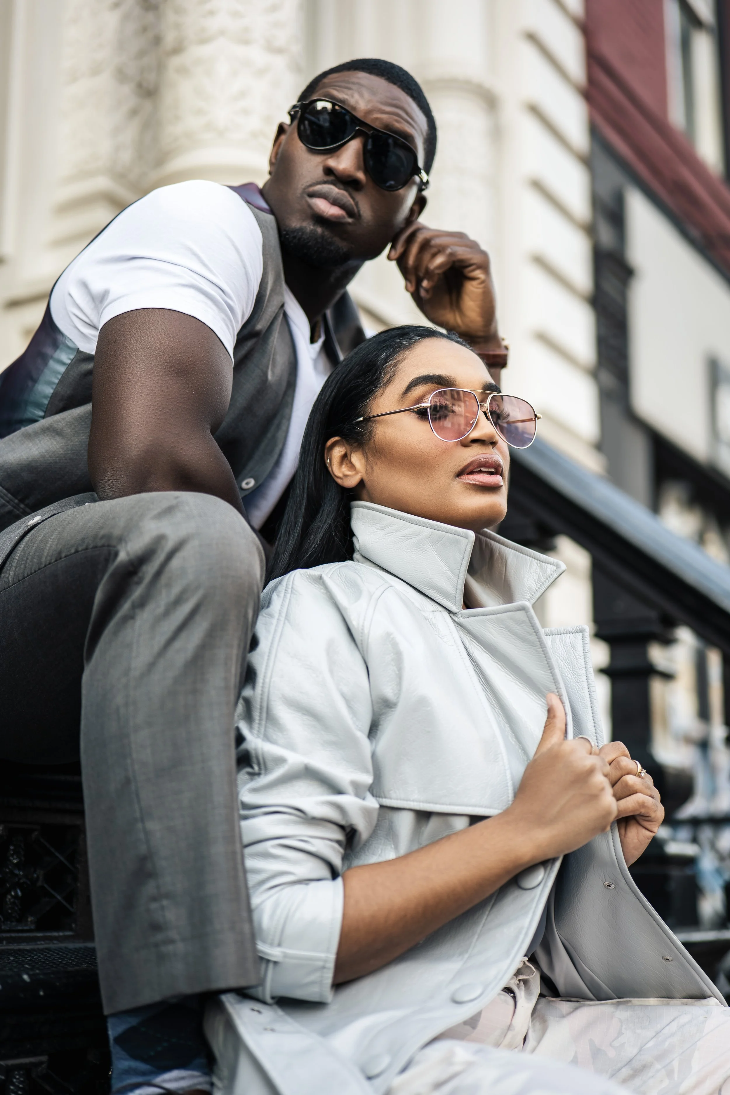 A young man and woman sitting on a bench outdoors in an urban area, both wearing sunglasses, with the man resting his chin on his hand and the woman with her hands clasped on her chest, looking thoughtfully into the distance.