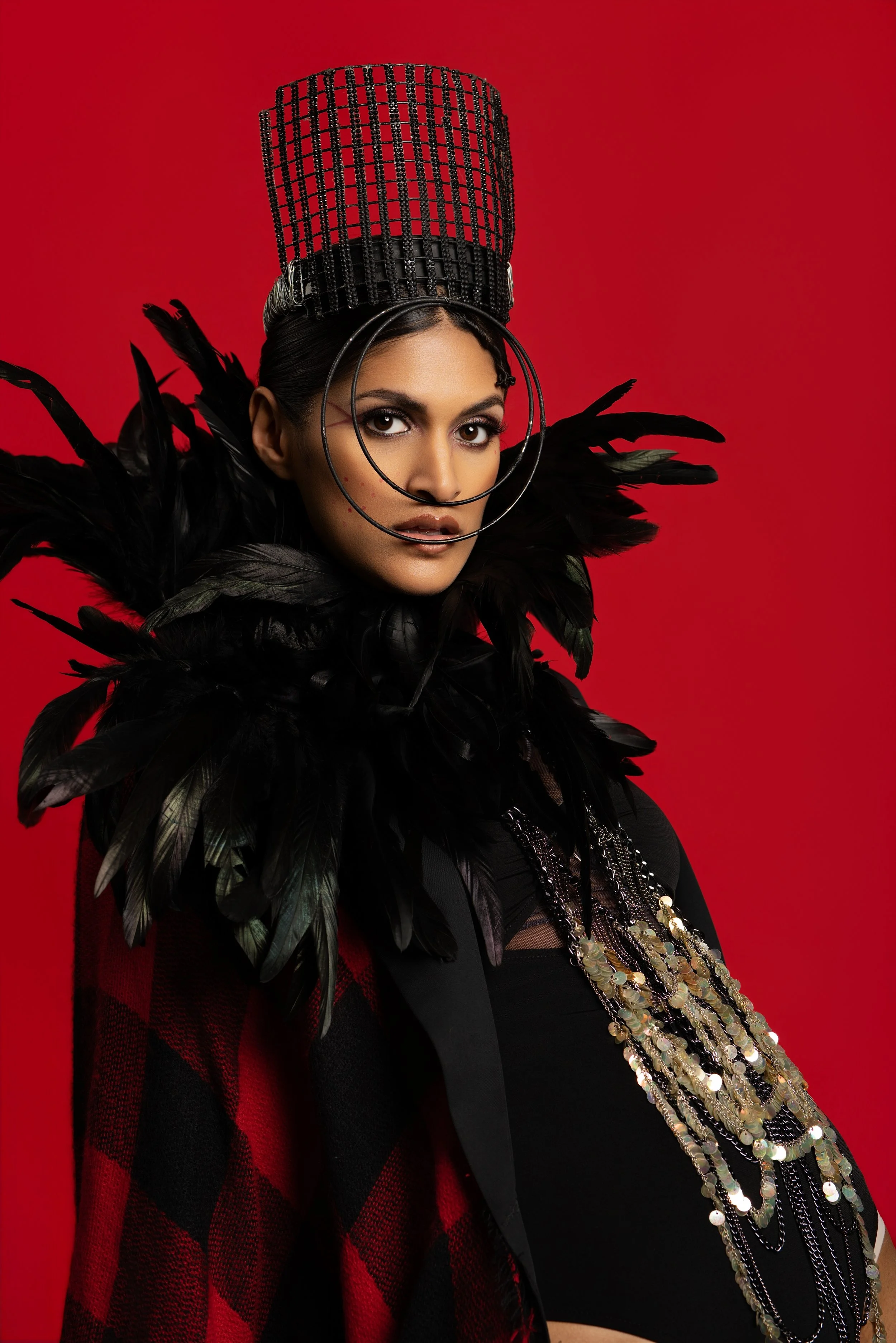 A woman wearing elaborate black feathered outfit and a tall, black beaded headdress standing against a red background.