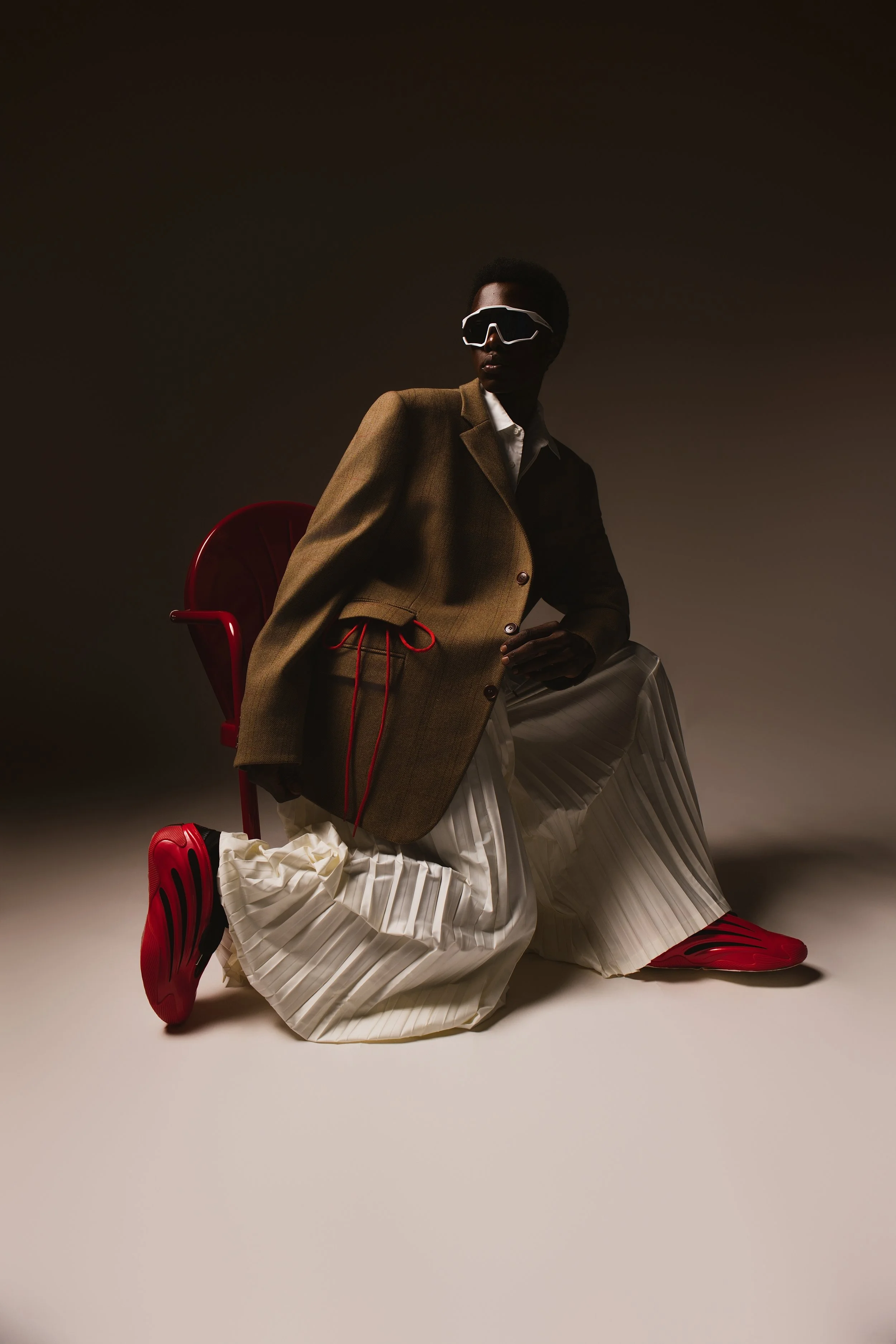 A fashion model wearing a brown blazer over a white shirt, with oversized white sunglasses, sitting on the floor with red sneakers, white pleated skirt, and a red chair in the background, against a dark background.