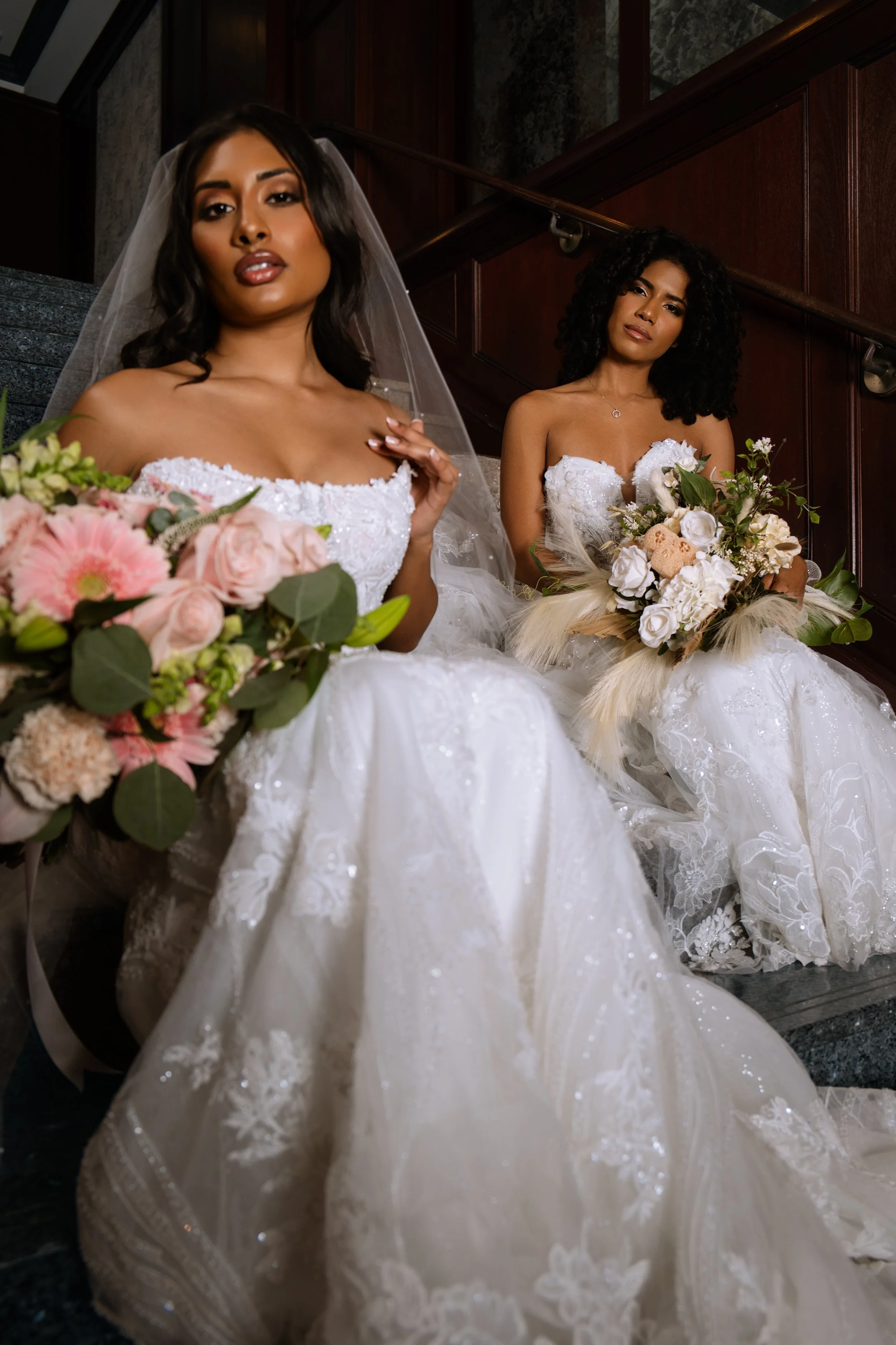 Two women in wedding dresses sitting on stairs, holding bouquets of flowers, with dark wood-paneled walls in the background.