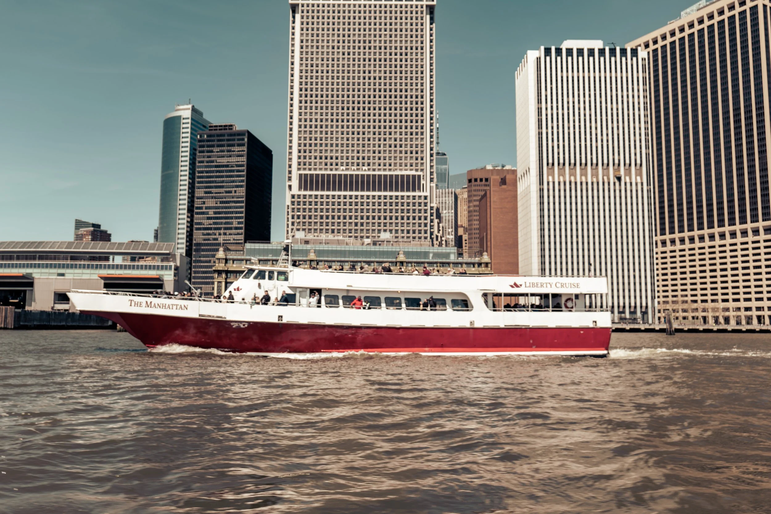 A red and white cruise boat named "The Manhattan" on the water with a city skyline in the background.