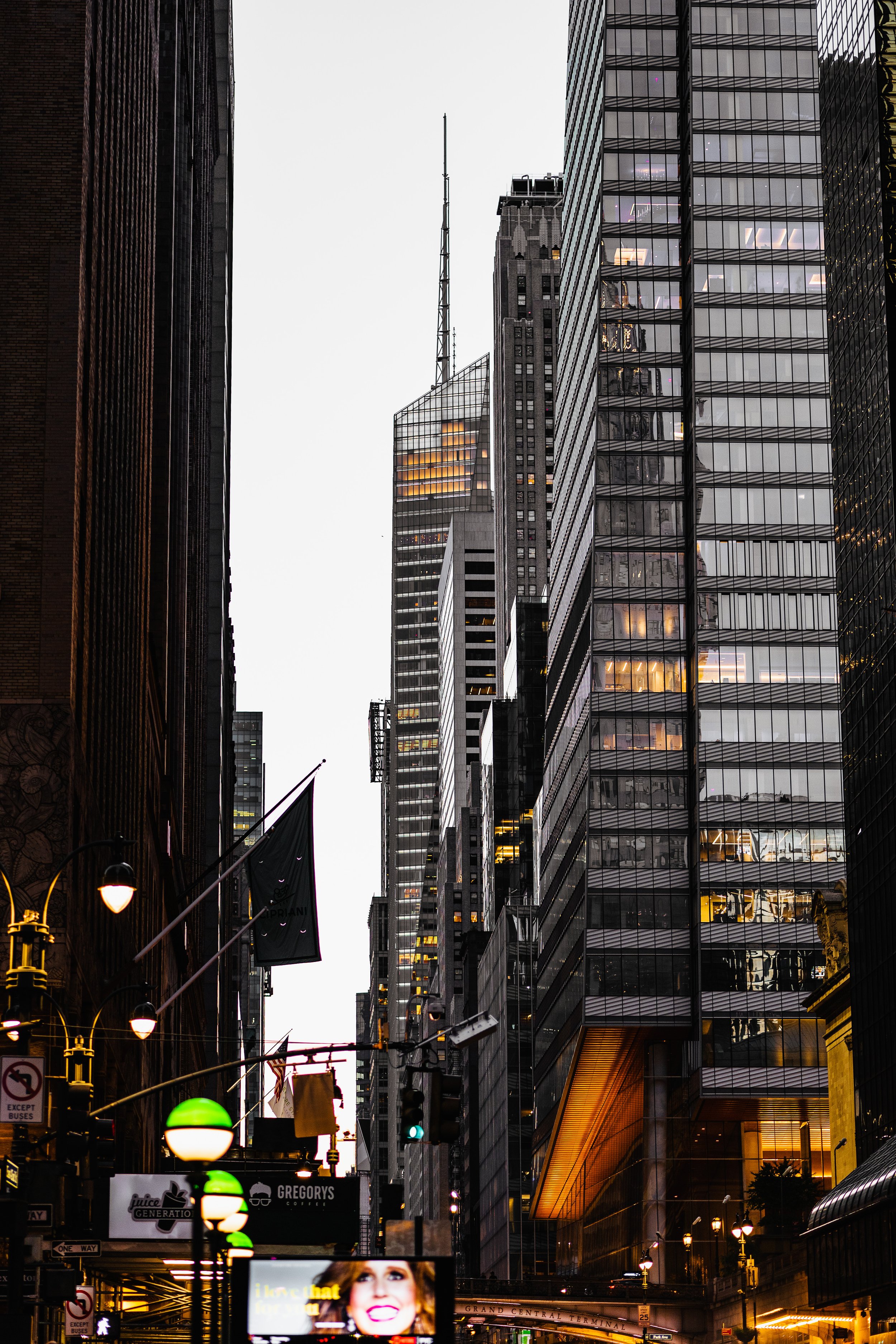 Urban street with tall skyscrapers, illuminated signs, and streetlights in a city setting.