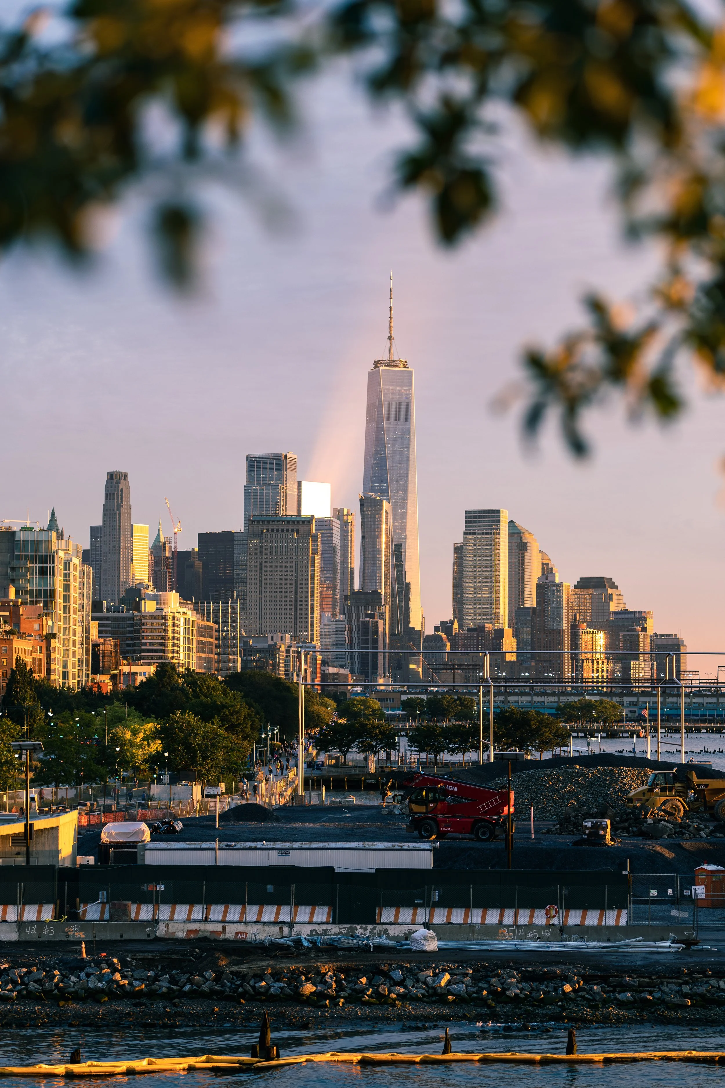 New York City skyline with One World Trade Center at sunset, viewed from a construction site in the foreground, framed by blurred tree branches.