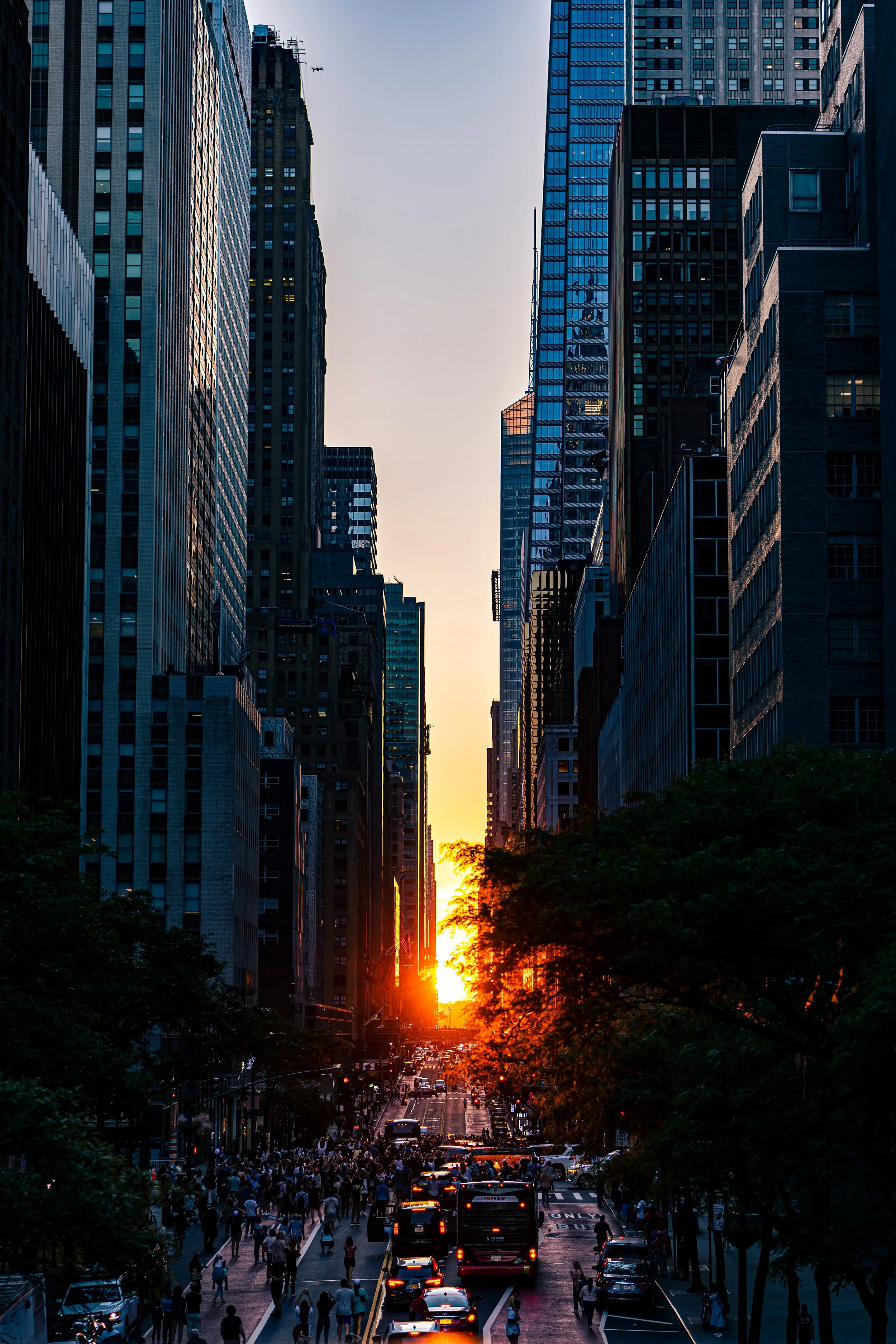 Sunset between skyscrapers in a city street with bustling traffic and pedestrians.