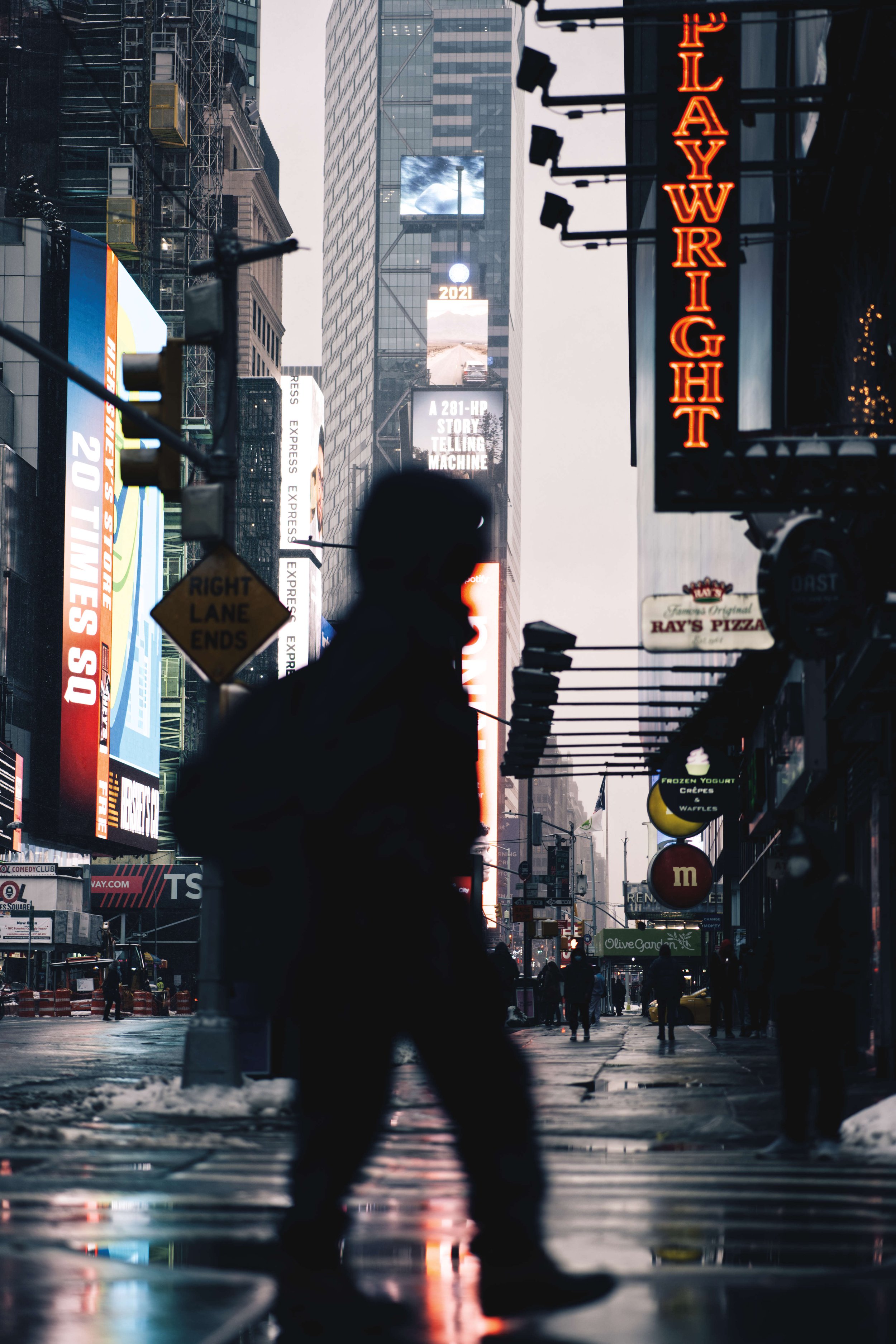 Silhouette of a person walking in Times Square, New York City, with bright neon signs, including "Playwright," and billboards in the background on a wet street.