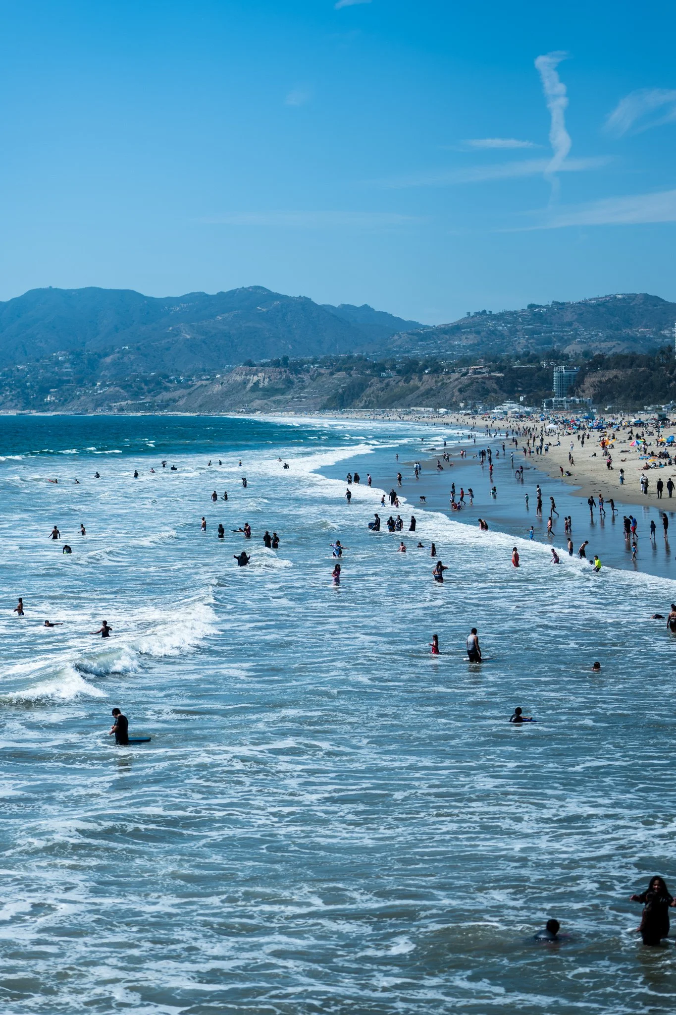 Beach with people swimming in ocean, sandy shore, mountains in background, clear blue sky.
